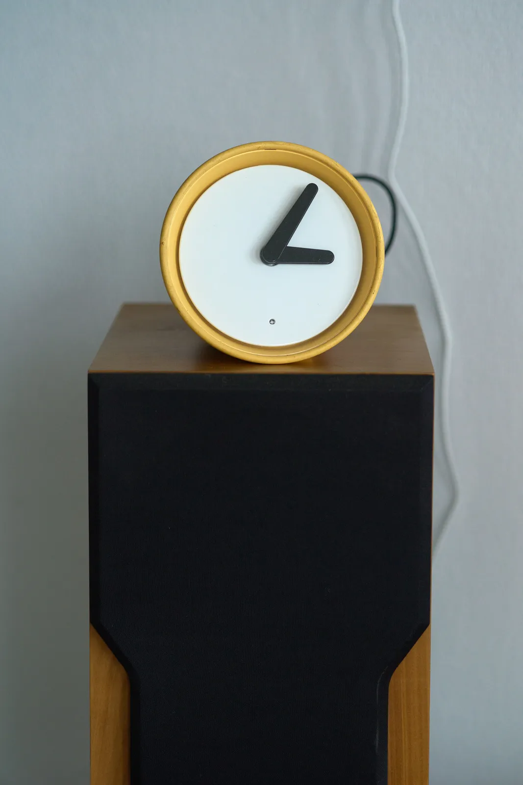 A yellow and white minimalist clock with black hands, on top of a designed speaker. It points at 5 past 3.