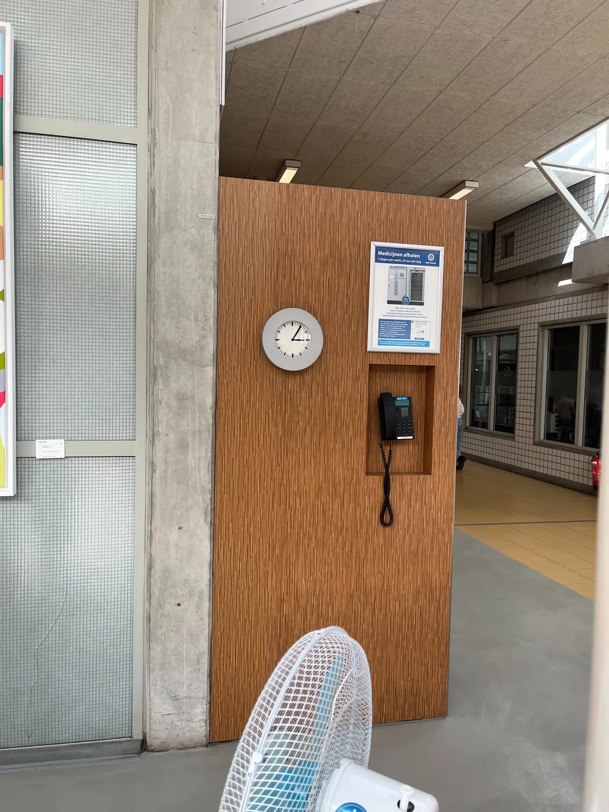 Inside a building. There’s a part of a fan visible. You can look past a wooden wall into a corridor. On this wooden wall there’s a clock. It points at 5 past 3