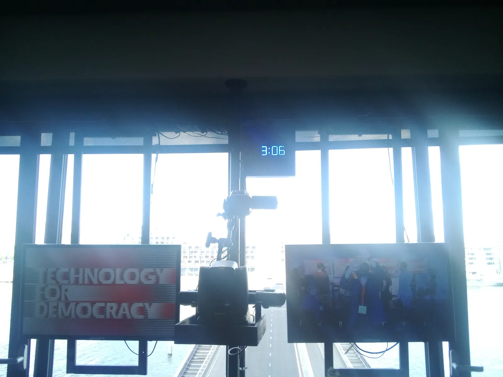 Looking down, through a window at a road. In front of the window there are two TV screens. One of them shows the text Technology for Democracy, and the other shows, a bit dark, the person who is taking this picture. Between the two screens there’s a digital clock that tells the time as 3:06