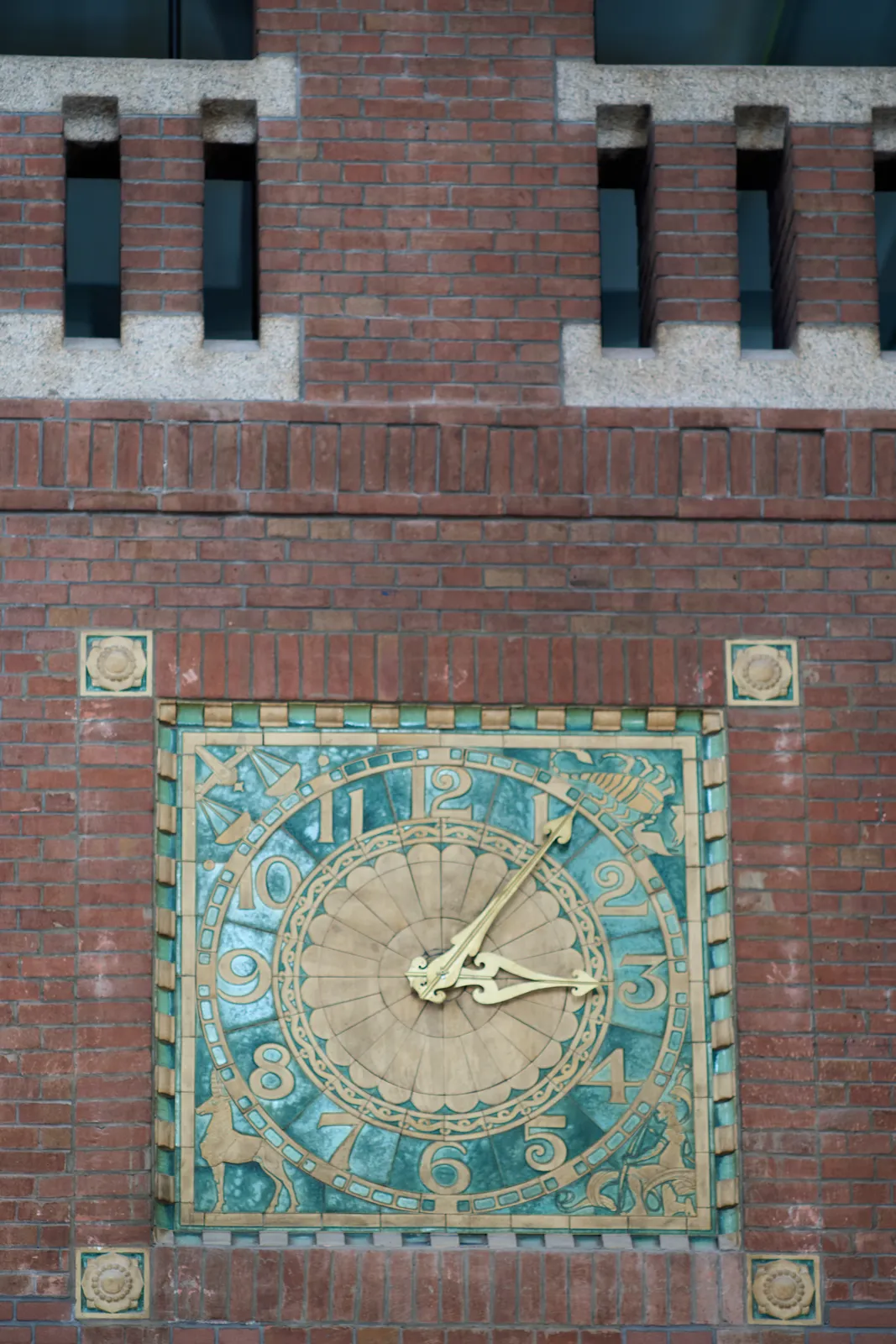 An ornamented clock, cemented into a brick wall. There are illustrations of a lobster, a unicorn, some scales and someone kneeling in the corners of the clock. The clock points at 6 minutes past three.