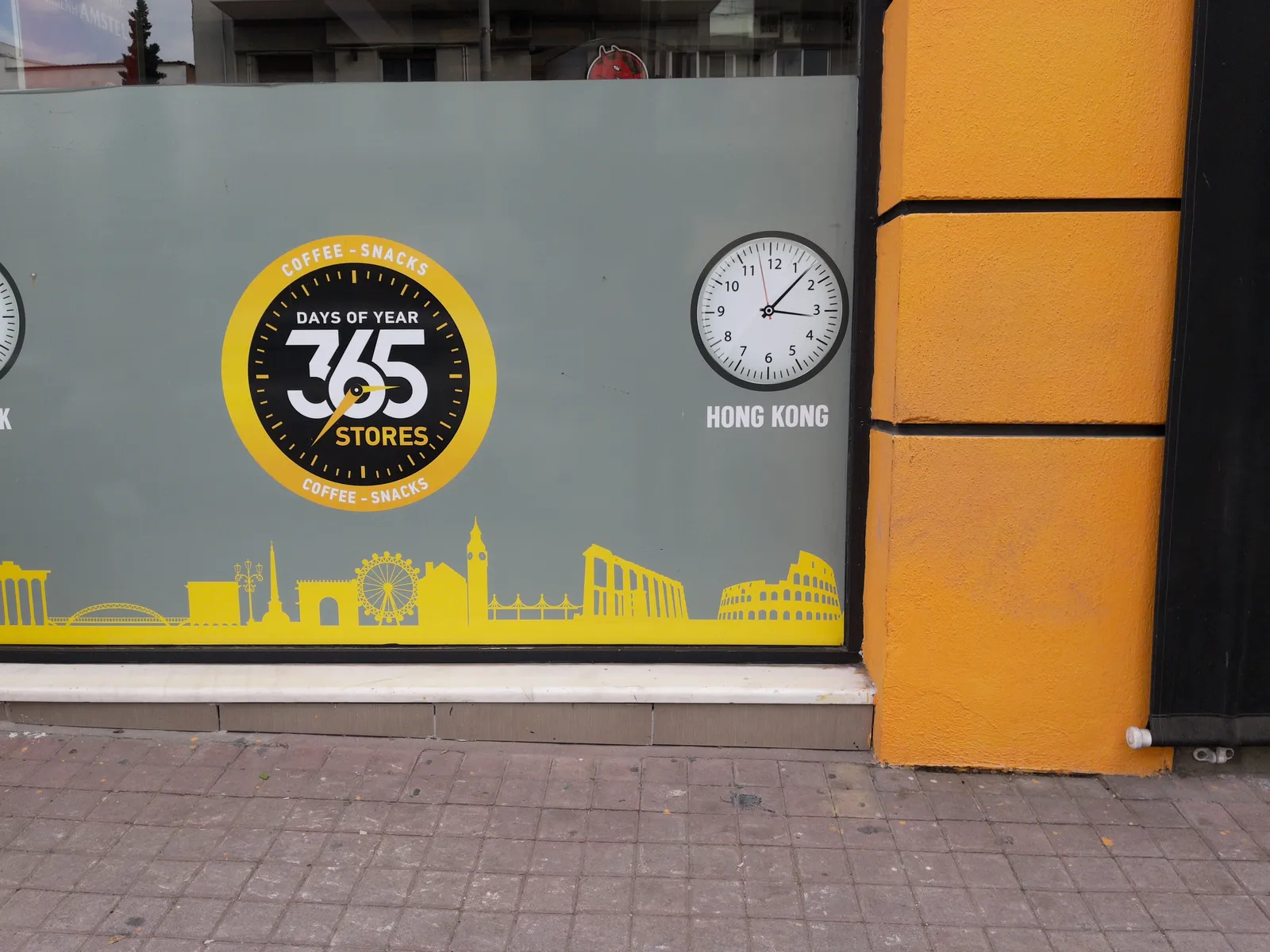 A store window, with all kinds of international landmark buildings printed on it, like the Colosseum, and the Big Ben. There are also two clocks printed on it. Below one of them it says Hong Kong. This one points at 7 past three. The other clock is the logo of the shop. It says coffee-snacks, days of the year 365 stores on its face. This clock points at 36 minutes past 2.