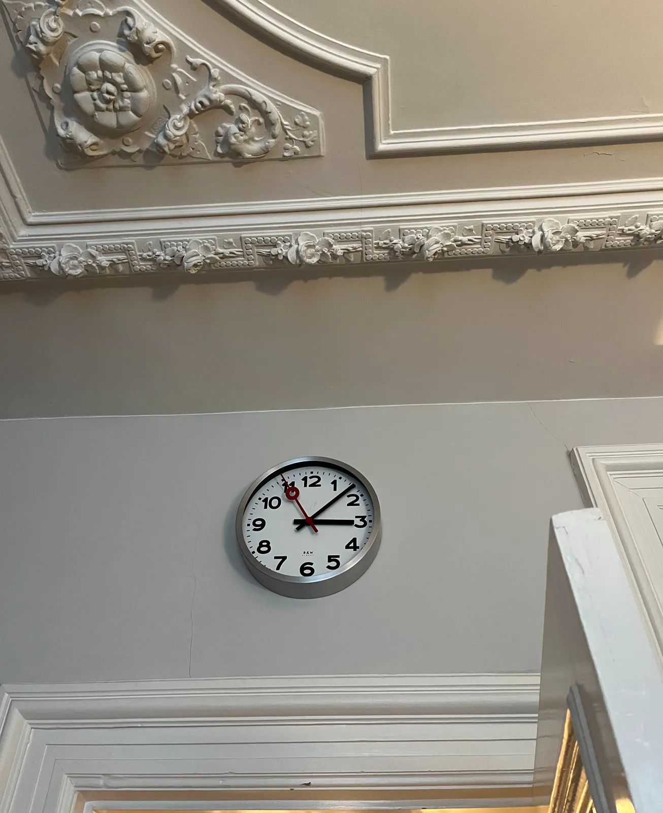 A richly ornamented ceiling, and an open, multilayered door. Above the door is a modernist clock that points at 7 minutes past 3.