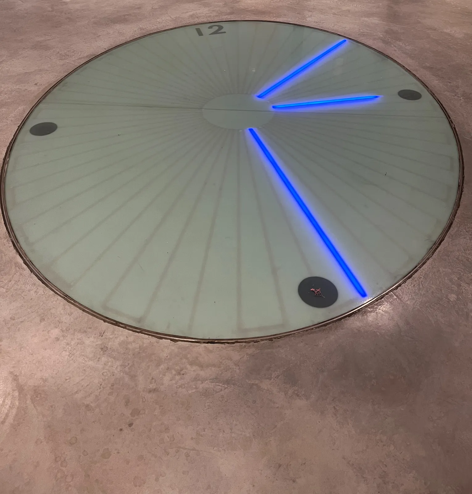 A clock built into the floor with hands made out of blue/purple neon. It points at 7 minutes past 3.