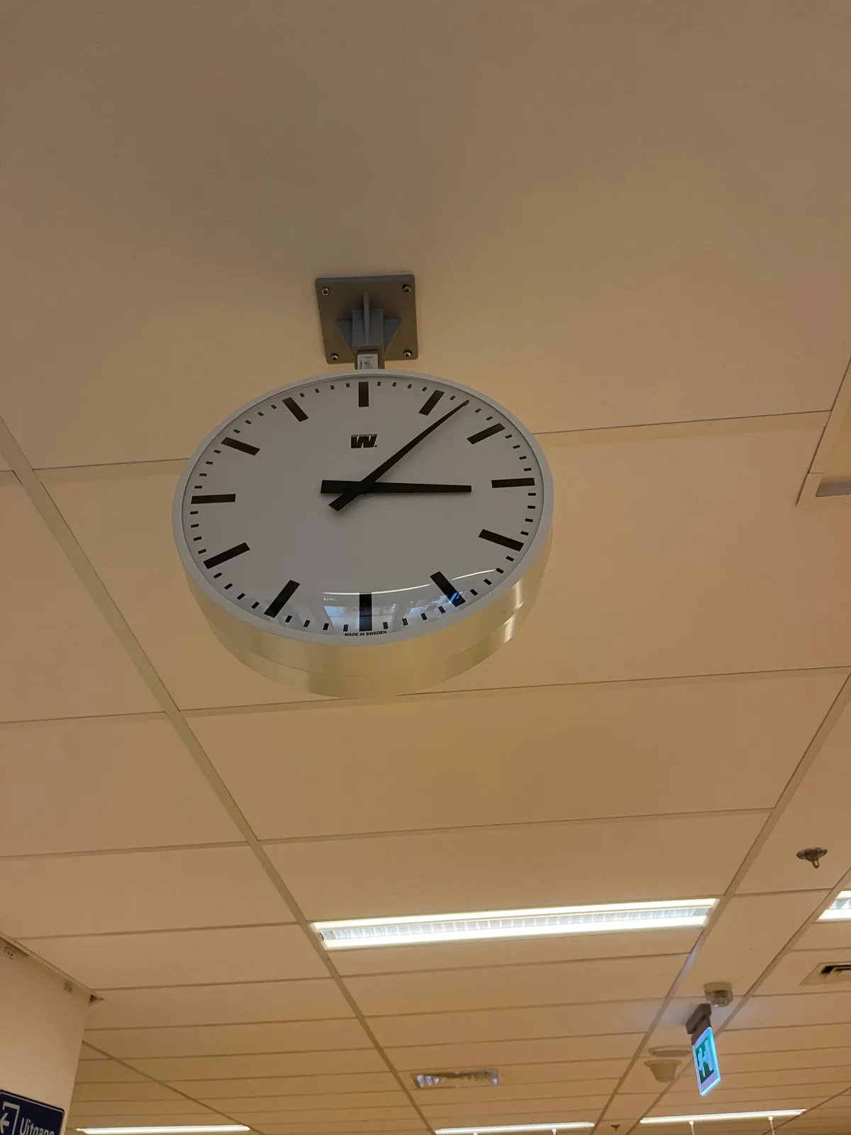 A white, modernist clock with pointy black hands, attached to a dropped ceiling. It points at 7 minutes past 3.
