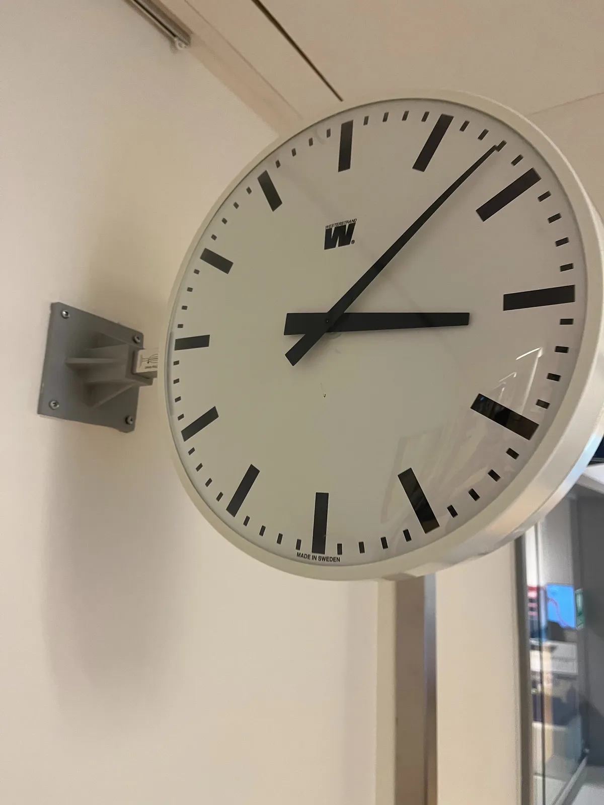 A white, modernist clock with pointy black hands, attached to a white wall. It points at 7 minutes past 3.