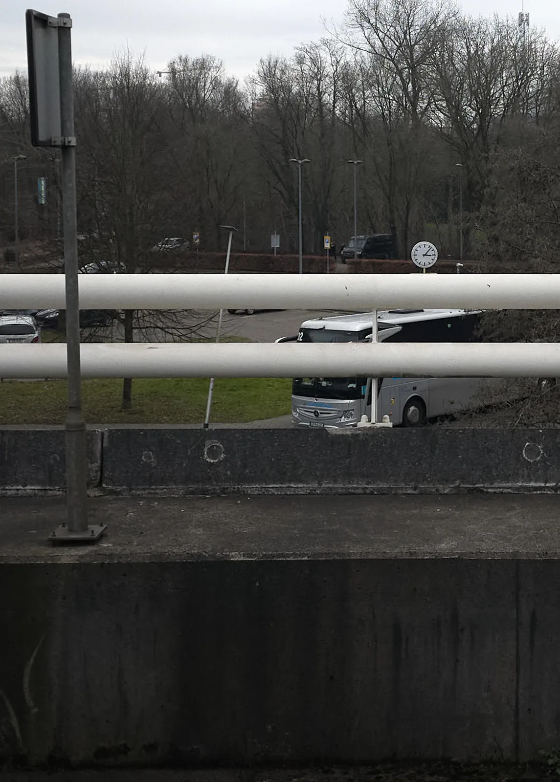 Peeking over the railing of a bridge, at a car parking. A large bus is parked. There’s a clock on the parking and it points at 7 minutes past 3.