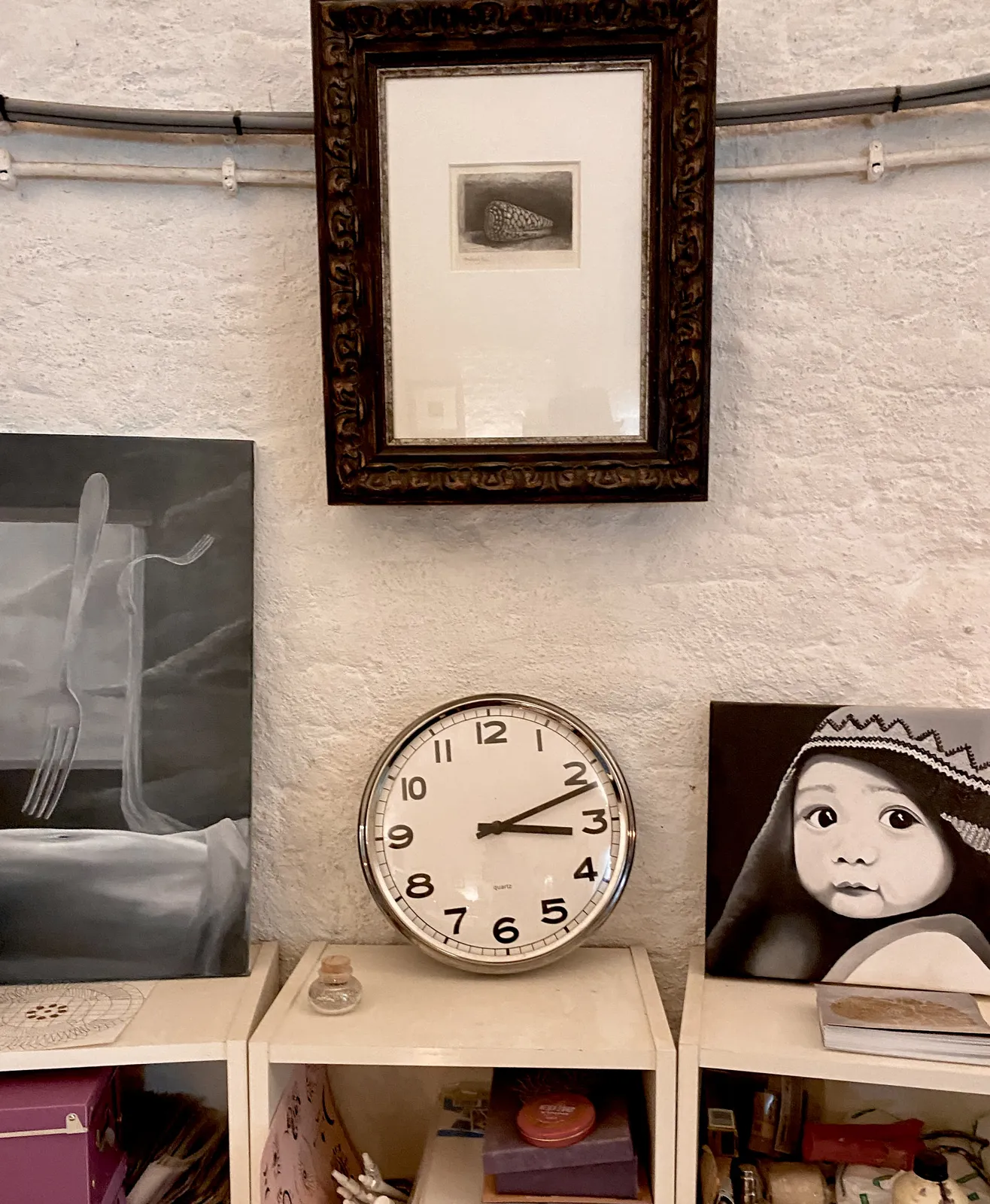 A few works of art, all black and white, and a clock. There’s a small drawing of a shell in a pretty big frame.  A painting of a floating fork, and a painting of a child’s face. The clock, which is black and white as well, points at 11 minutes past 3.