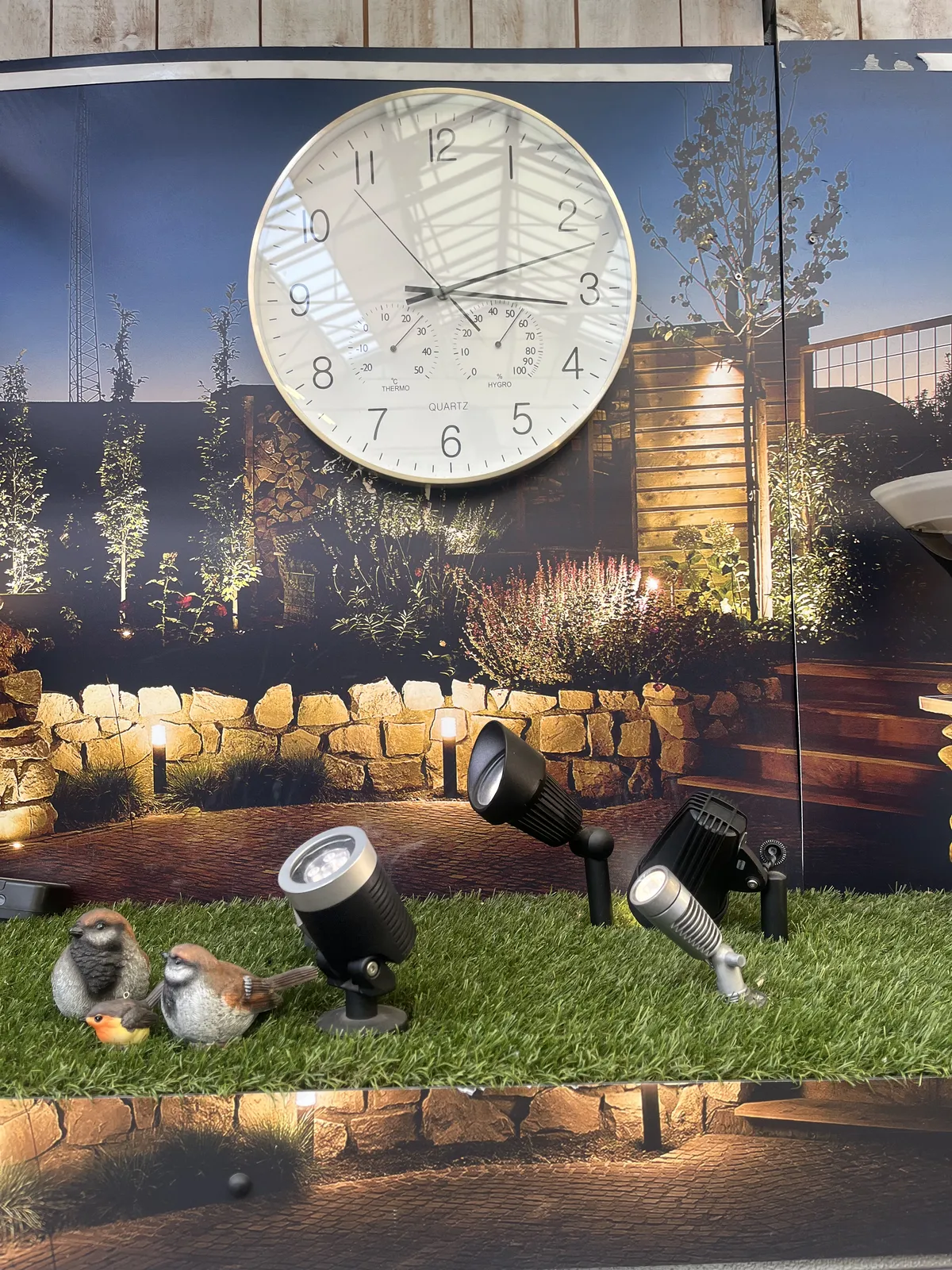 Fake birds are set on a piece of fake grass, next to some lamps. Behind it is a poster of a garden, with a working wall clock on it. This clock points at 12 minutes past three.