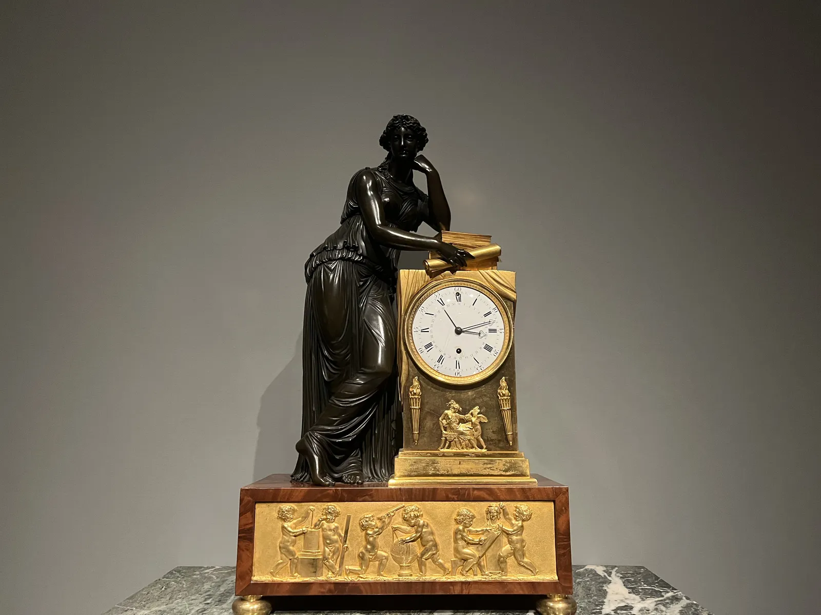 A black statue of a woman leans against a golden clock. The clock points at 12 minutes past 3.