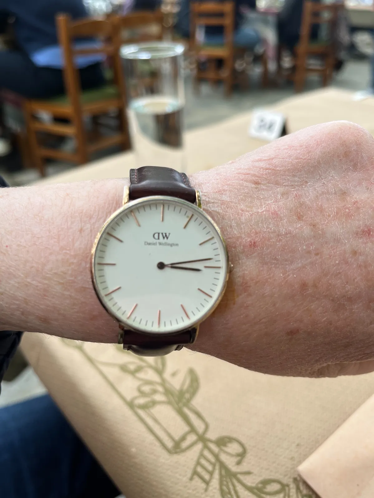 Someone is holding up their arm to show the watch on their wrist. In the background you can see the charisteristic, uncomfortable wooden chairs of Greek tavernas. The wrist watch points at 13 minutes past 3. 