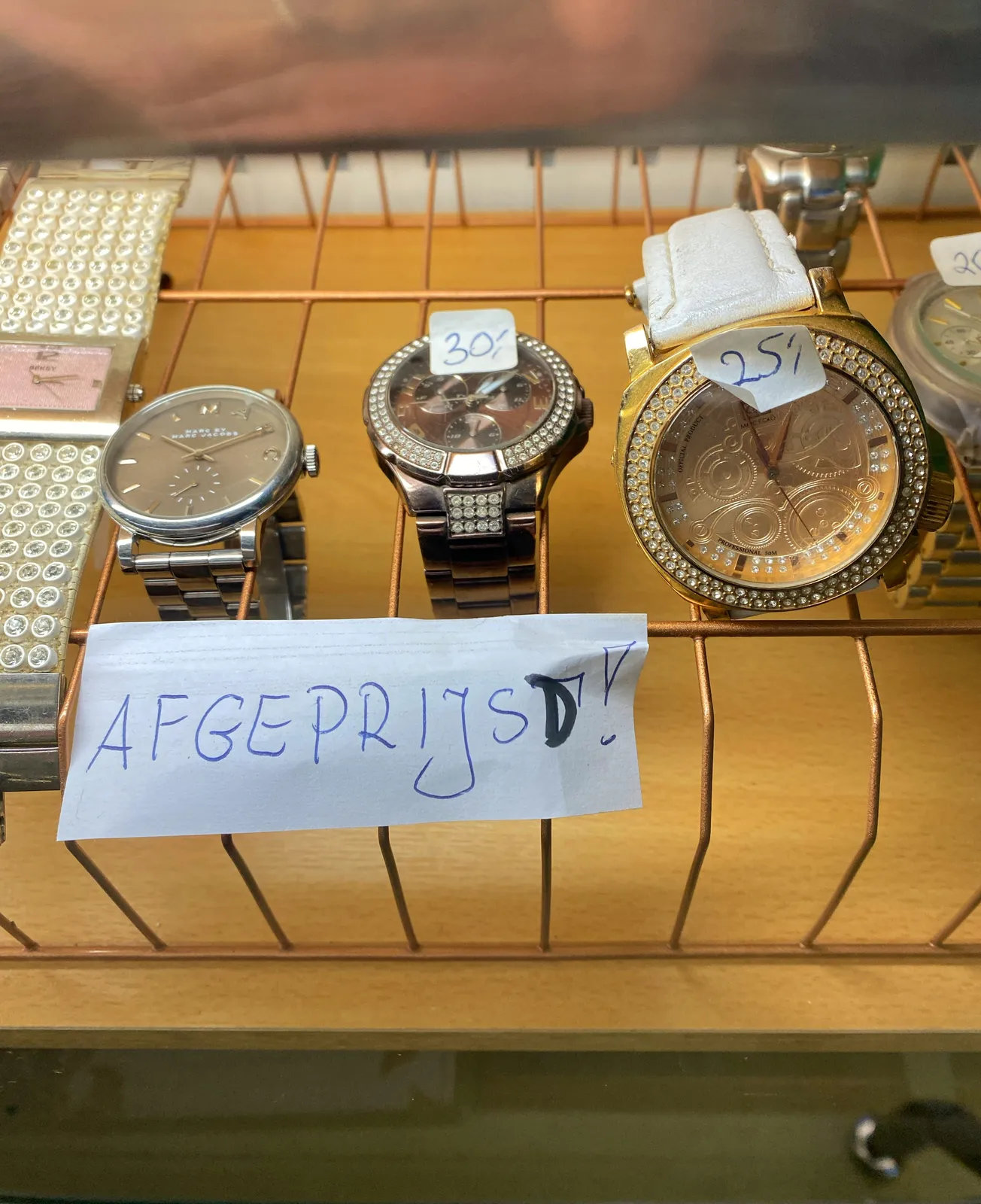 There are four different, shiny watches in this picture. The one on the left, with a wrist band made from diamonds (probably fake, looking at the low price of these watches) points at 13 minutes past 3. The one next to it, which is the least blingy, points at 11 past 10. Then, the next watch with some (probably fake) diamonds on the ring around the face points at 8 minutes past one. And finally, the clock on the right, with lots of diamonds on and around its golden face points at 2 minutes to one. There’s a handwritten sign that says all these watches are on discount. One of them is 25, another is 30.