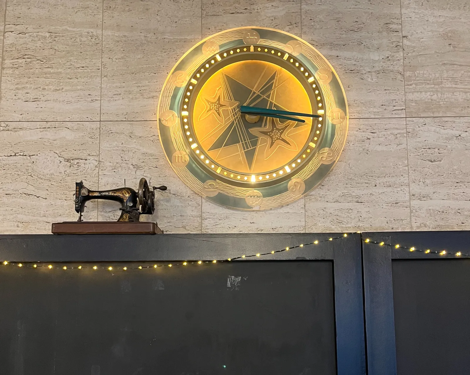 A wall clock, nicely lit. It has the zodiac signs as hour indicators. It points at twins past twins, which translates as a quarter past 3. Next to this clock there’s a very old, black, mechanical sewing machine.