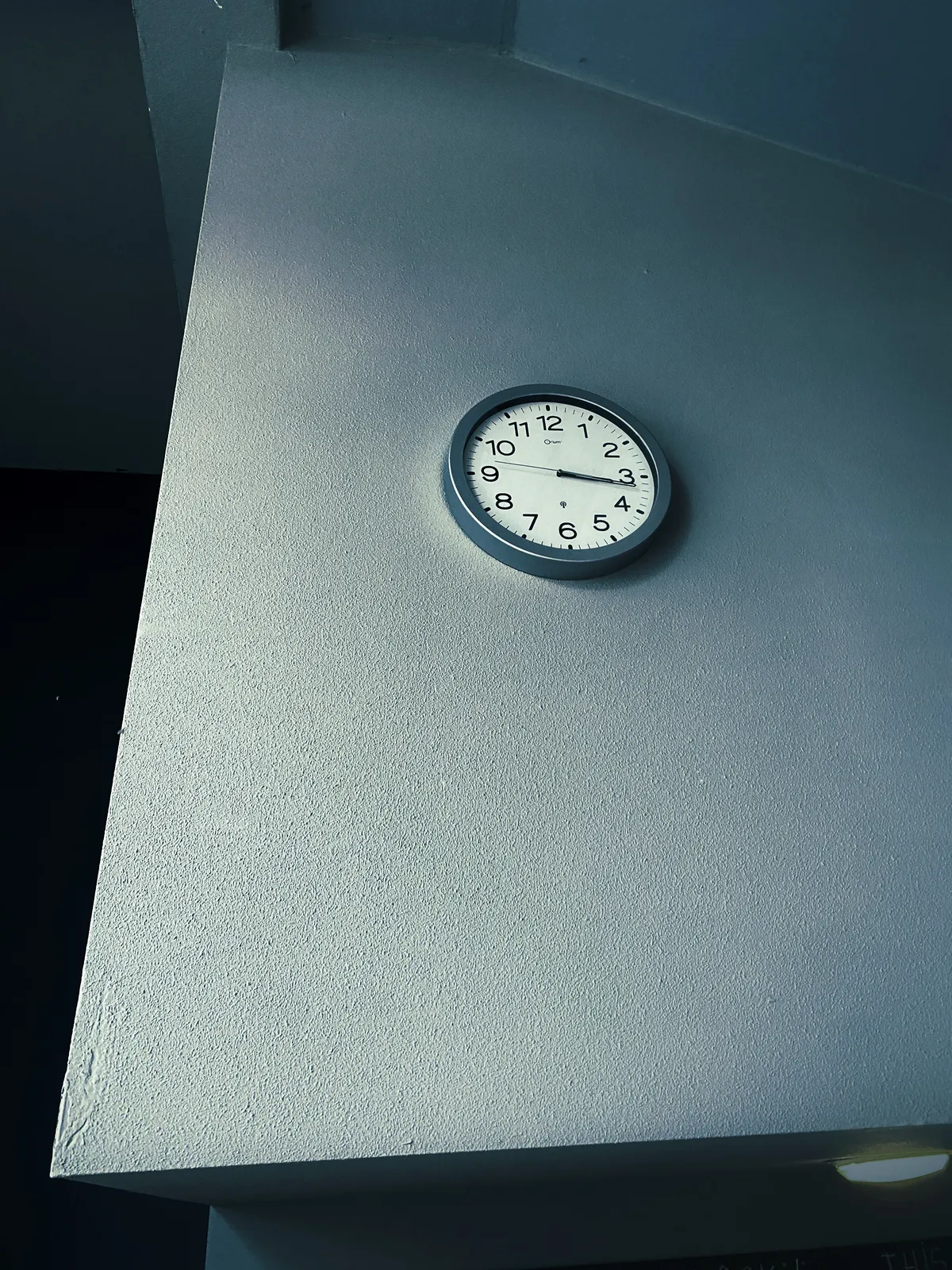 A clock in the middle of a seemingly triangular wall. The whole picture is blueish. The clock points at 16 past 3.