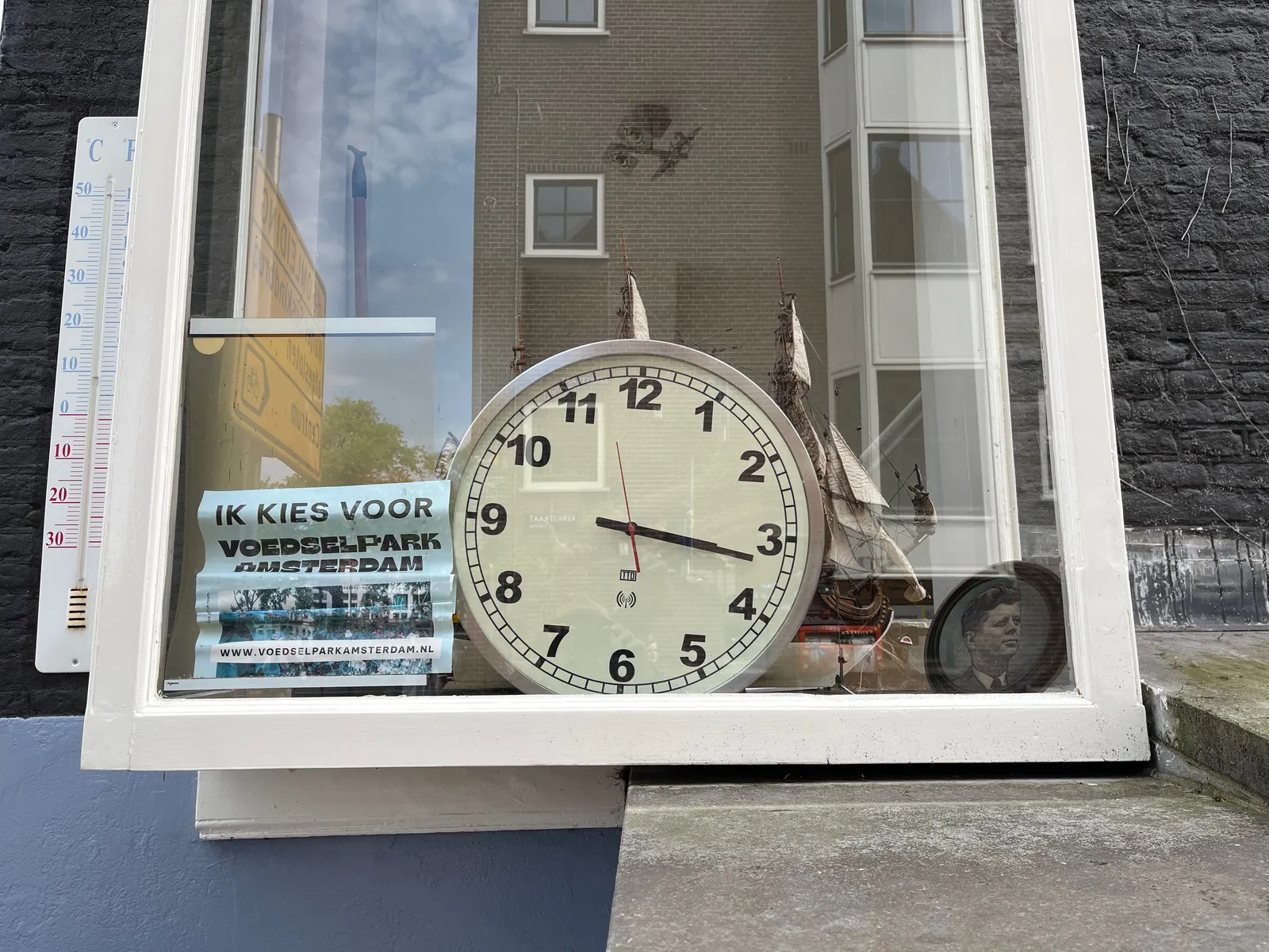 Lots to see in this image. In the reflection of the window there’s an apartment building and a yellow sign indicating a detour for bicycles. Next to the window there’s a thermometer: It’s unclear how warm it is. In the window there’s a portrait of John F. Kennedy, a poster promoting a food park in Amsterdam, a model of an old ship, and a clock. The clock points at 16 past 3.