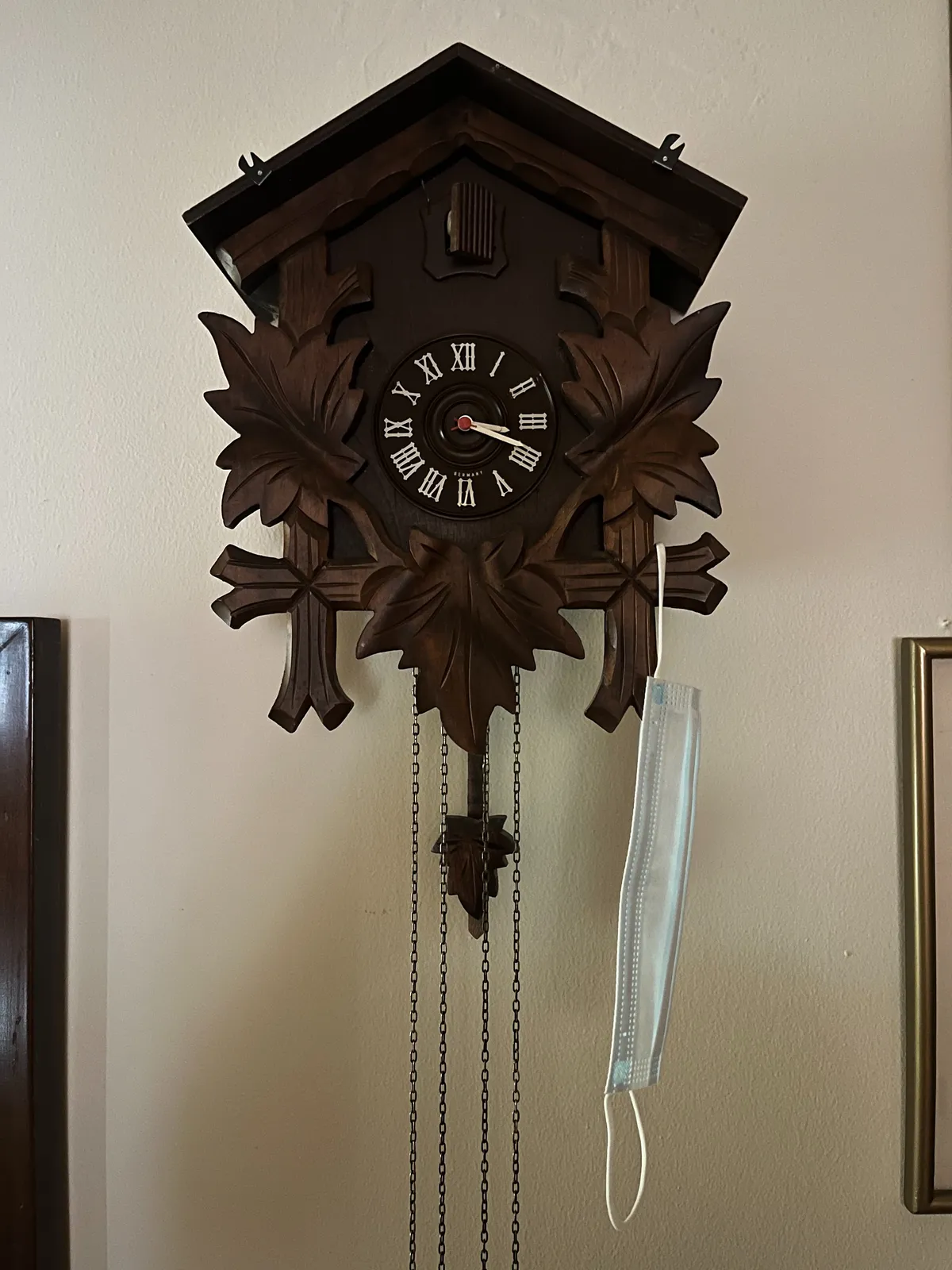 A cuckoo’s clock with a face mask hanging from one of its ornaments. It points at 18 minutes past 3