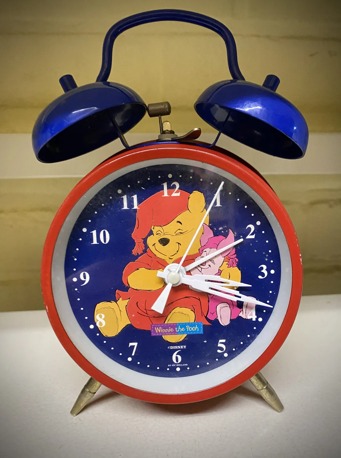 An old fashioned, red and blue mechanical alarm clock, the one with two bells on top. The bells are blue. This clock has Winnie the Pooh printed on its face. It points at 18 minutes past 3.