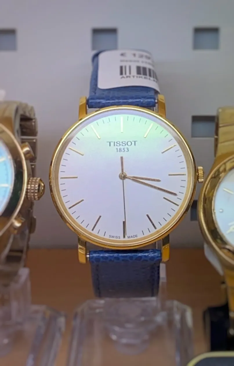 A simple wrist watch with a blue band, a white face, and gold details and a golden frame. On its face it says Tissot 1853, Swiss Made. It points at 18 minutes past 3.