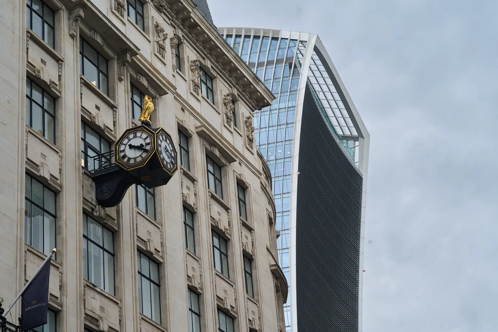 A clock attached to a high, white, rather old facade. Behind this building there’s a very modern sky scraper. The clock is black, with golden hands and indicators, and with a golden bird sitting on top of it. The clock, not the bird, points at 19 minutes past three. 