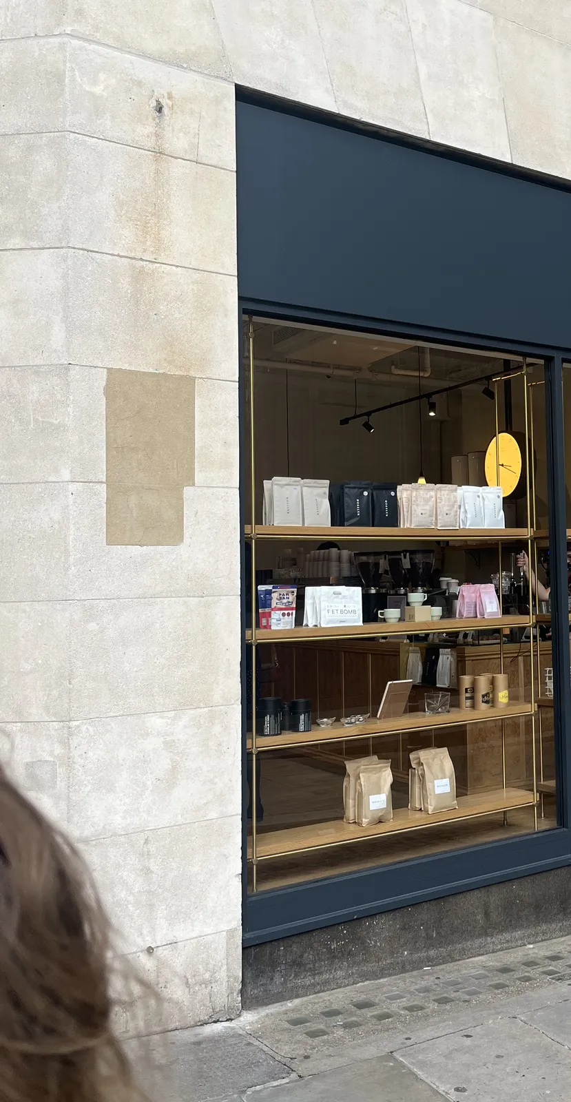 Peeking into a store, there are minimalist plastic bags in the window. Behind these bags, inside the store, a minimalist clock can be seen that points at 19 minutes past three.