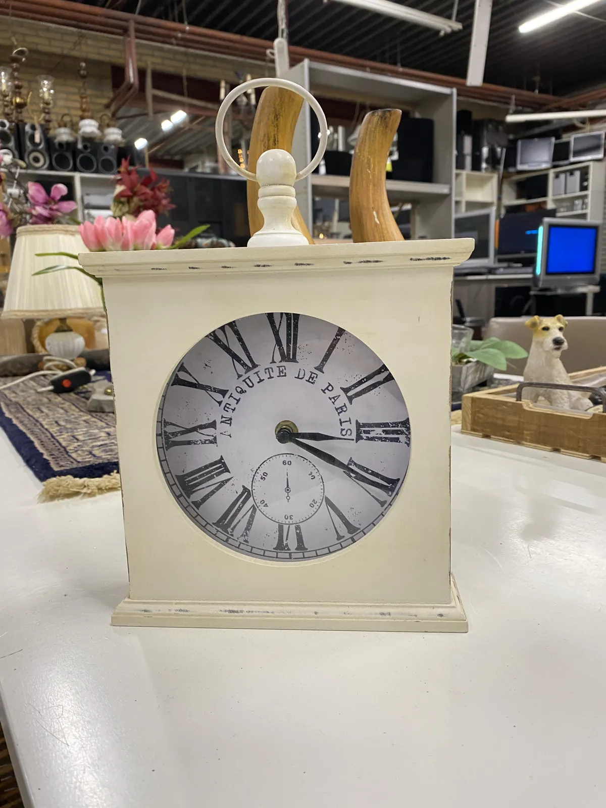 All kinds of stuff for sale in this large thrift shop. In the foreground there’s a white table clock with the words Antique de Paris printed on its face. It has roman numerals, printed as if its old. There’s also a fake dial printed on its face. The clock seems to work though and it points at 20 past 3.