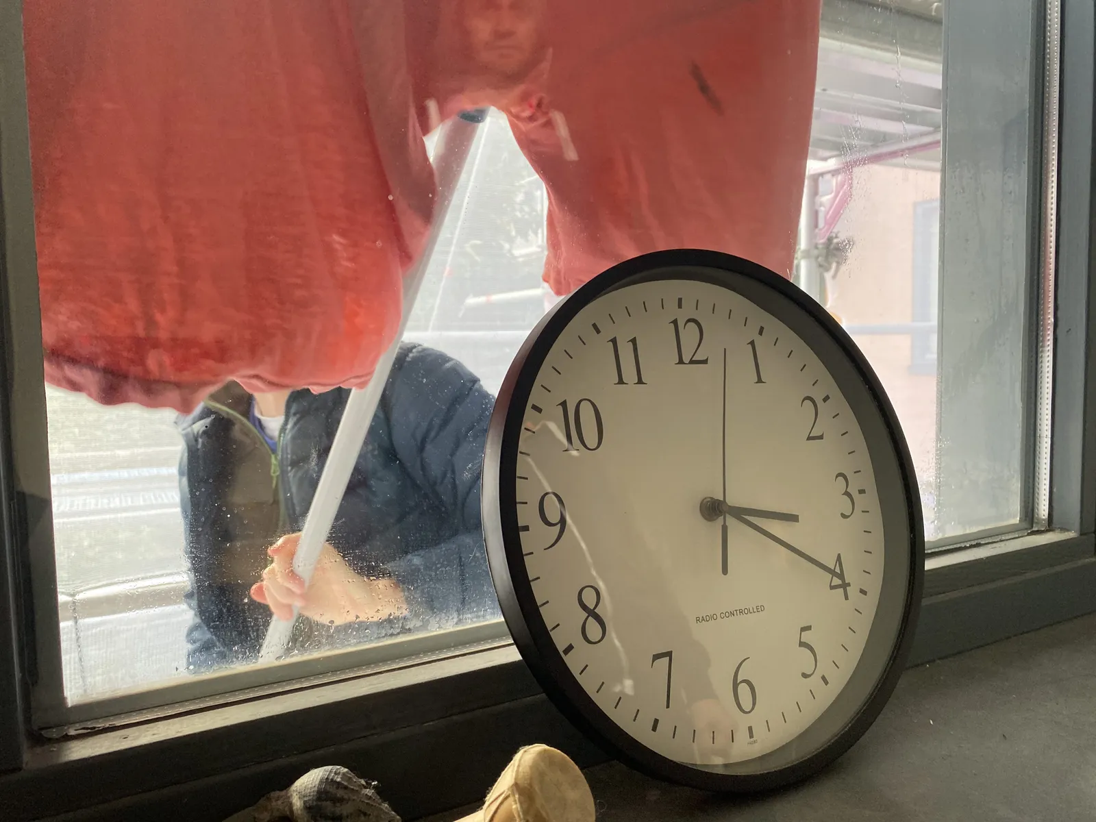 It looks like someone is cleaning the window on the outside with a red cloth. In front of the window there’s a clock. On it it is 20 past 3.