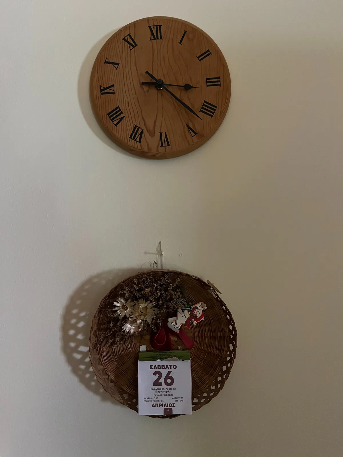 Below a wooden clock, attached to the wall, there’s basket with some flowers in it, and a calendar that says, in Greek that it is Saturday the 26th of April, which is almost certainly not true, right now. The wooden wall clock points at 22 minutes past 3.