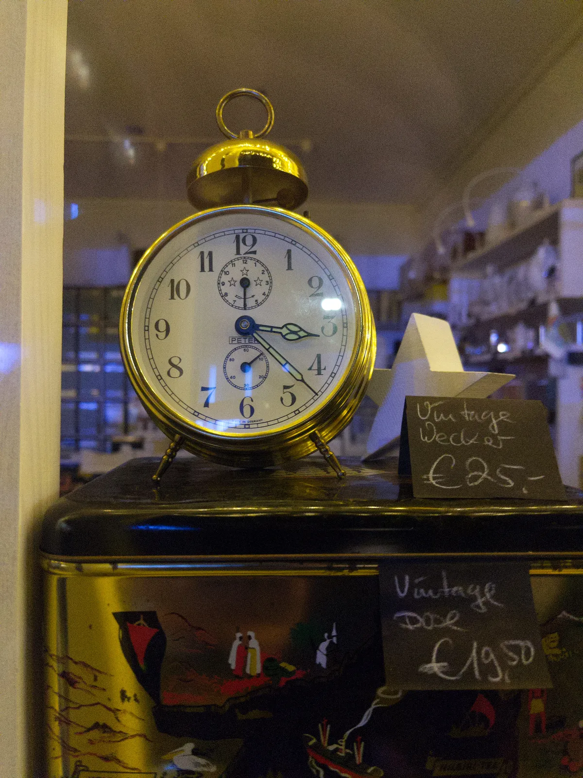 A vintage alarm clock with a single bell on top in a shop window. The clock points at 22 minutes past 3.