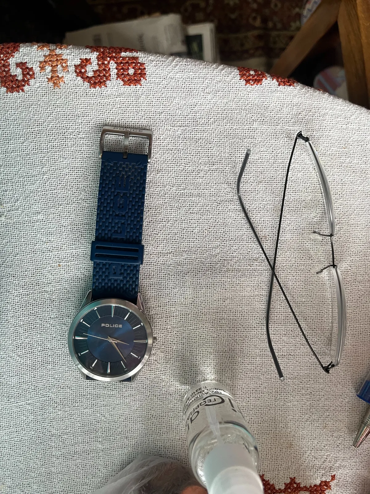 A little bottle with some transparent fluid in it, reading classes, and a wrist watch on an embroidered table cloth. The watch is blue, it bears the moniker Police, and it poinst at 23 minutes past three.