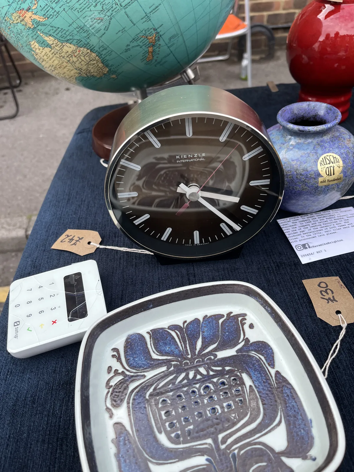 Things for sale. A plastic globe, a vase, and a plate. There’s also a small point of sale terminal. And there’s a clock for sale for 42 pounds. It points at 23 minutes past three.