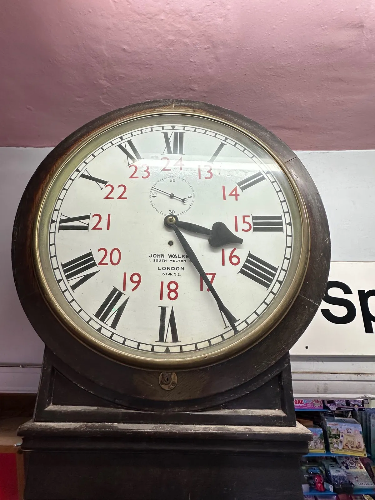 An old wooden clock. It has the hours from 1 to 12 printed in black roman numberals, and the hours from 13 to 24 in smaller red numbers. It points at 25 past 3.