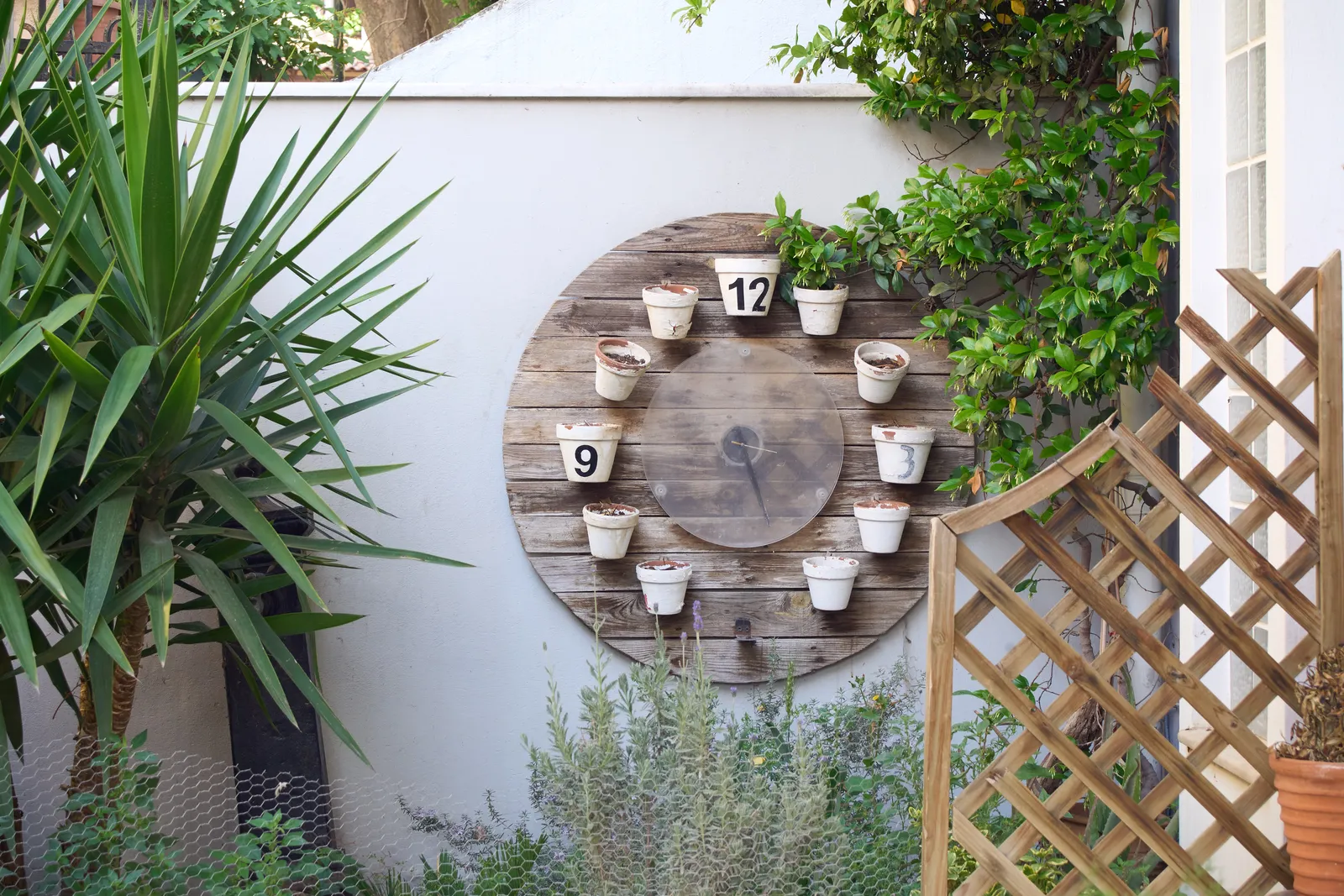 On a wall in a garden, surrounded by plants, is a large clock made from flowering pots. This clock points at 26 minutes past 3 