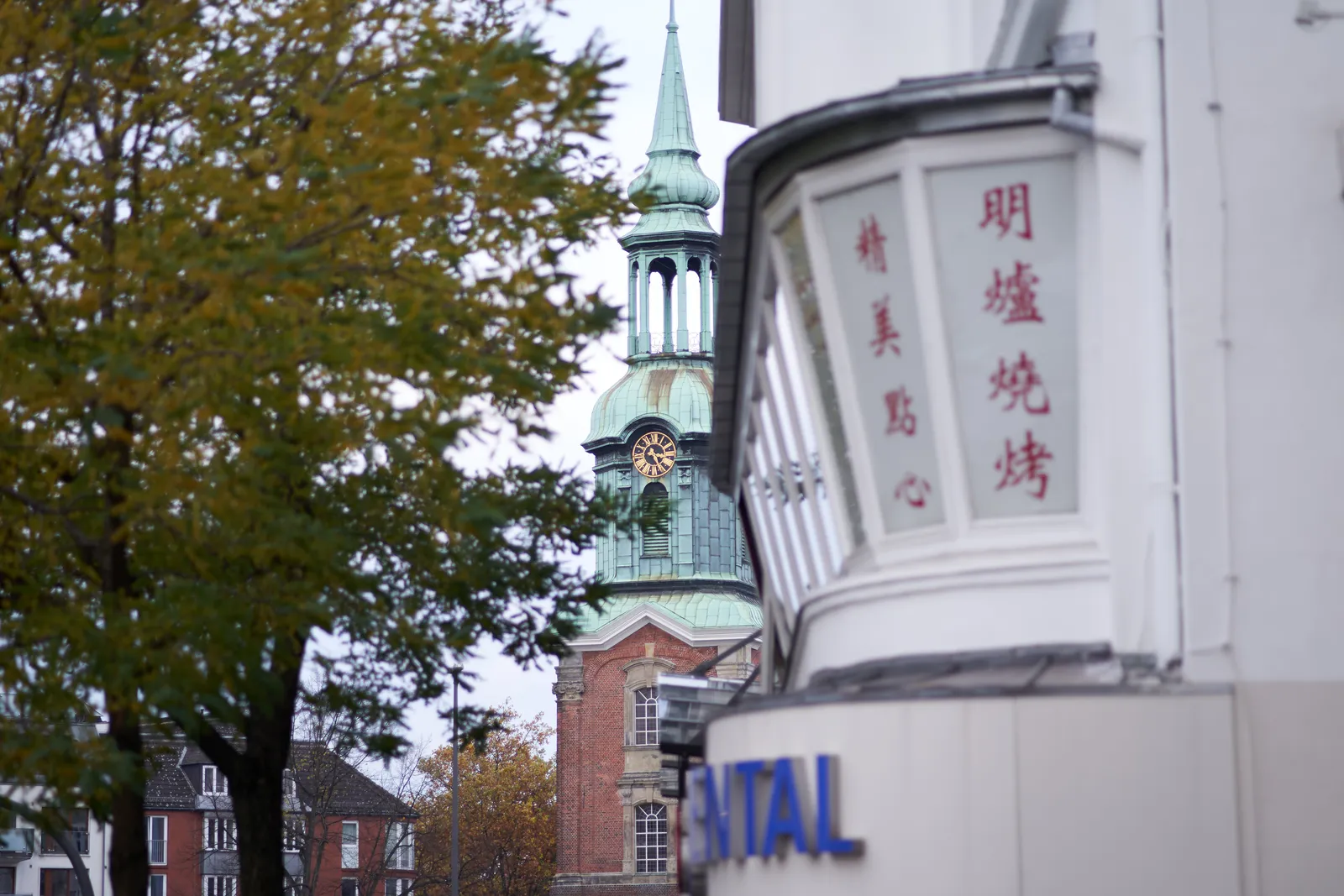Peeking through the branches of a tree, and past a Chinese restaurant at a clock on a church. It points at 26 past 3.