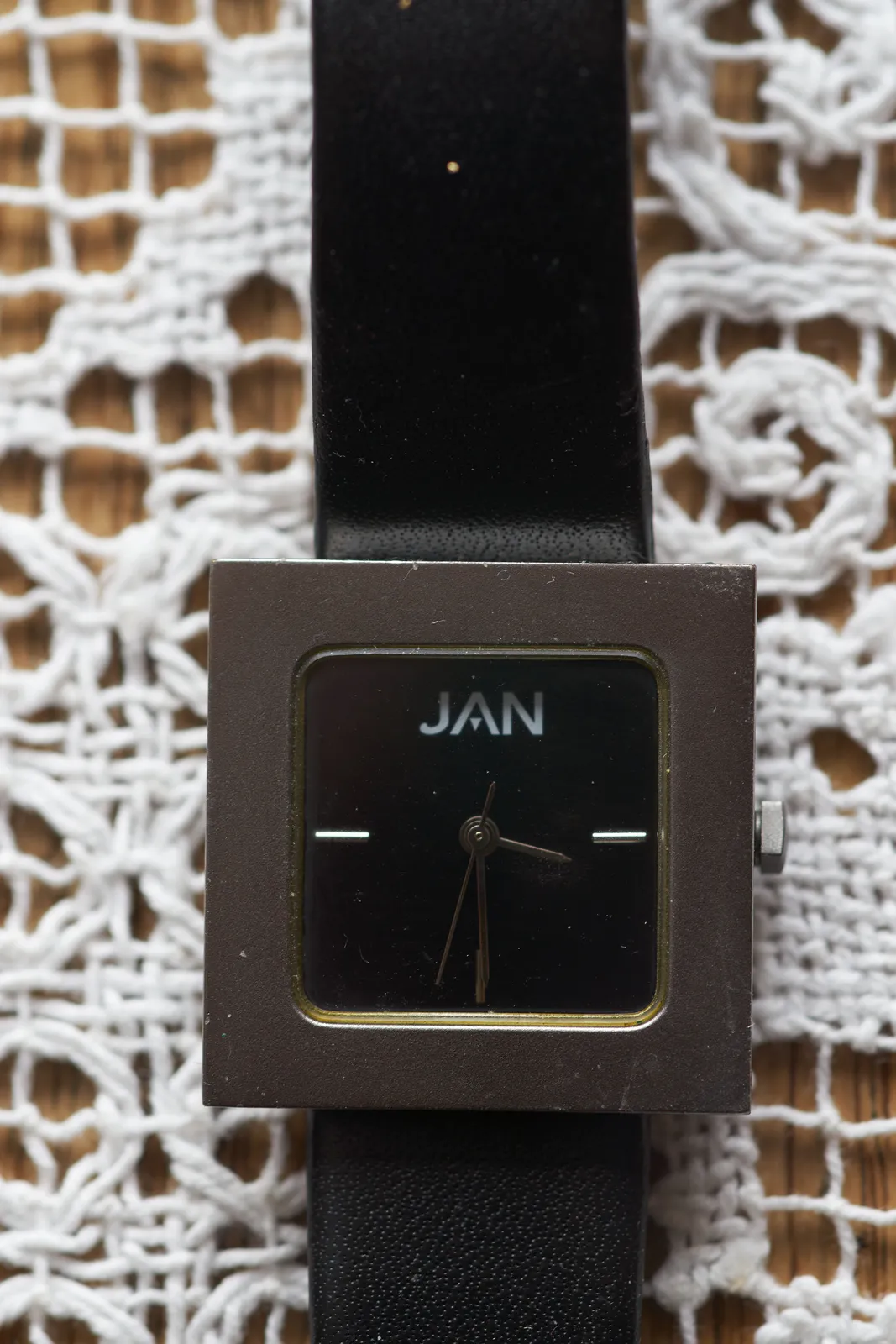 This minimalist, square, black and silver watch with the letters JAN on it, on a crocheted white fabric background points at 29 past three. 