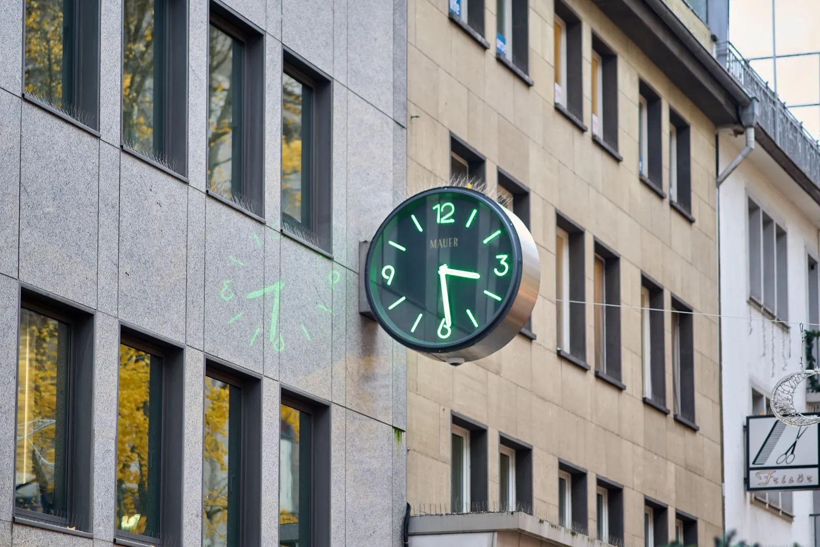 A black clock, with green, lit hands, attached to the facade of a building. The lit hands are also mirrored in the shiny facade. The clock points at 29 minutes past 3.