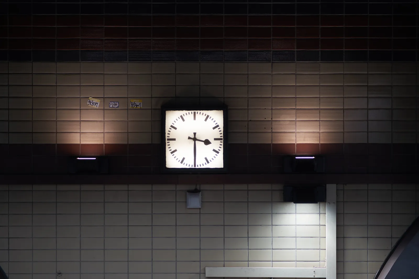 A clock on a tiled wall. The tiles are white and brown and there are some stickers on it. The clock points at half past 3.