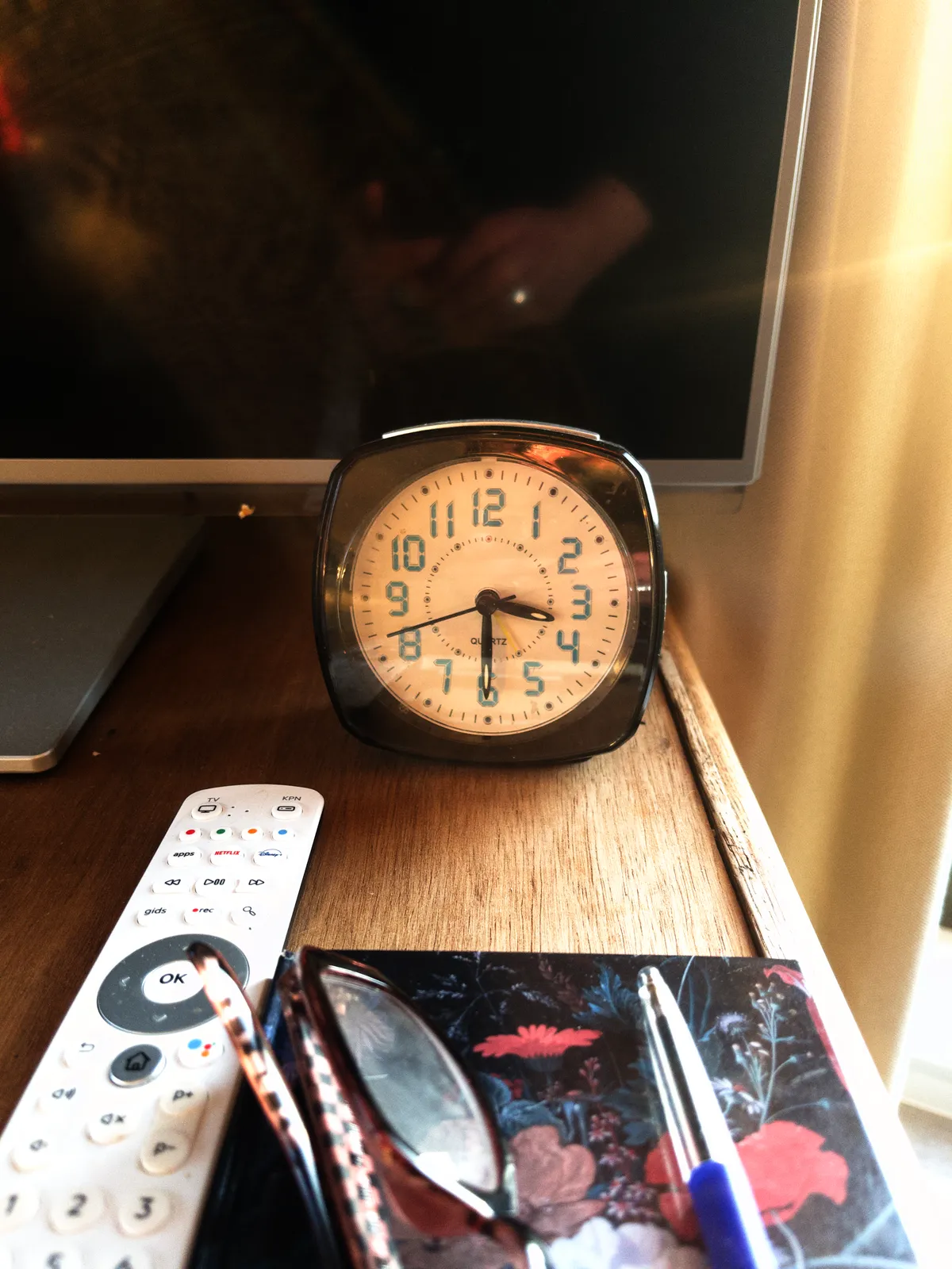 A small alarm clock in front of a tv-screen. There are a remote control, a pen and some glasses next to it. The clock points at half past three.