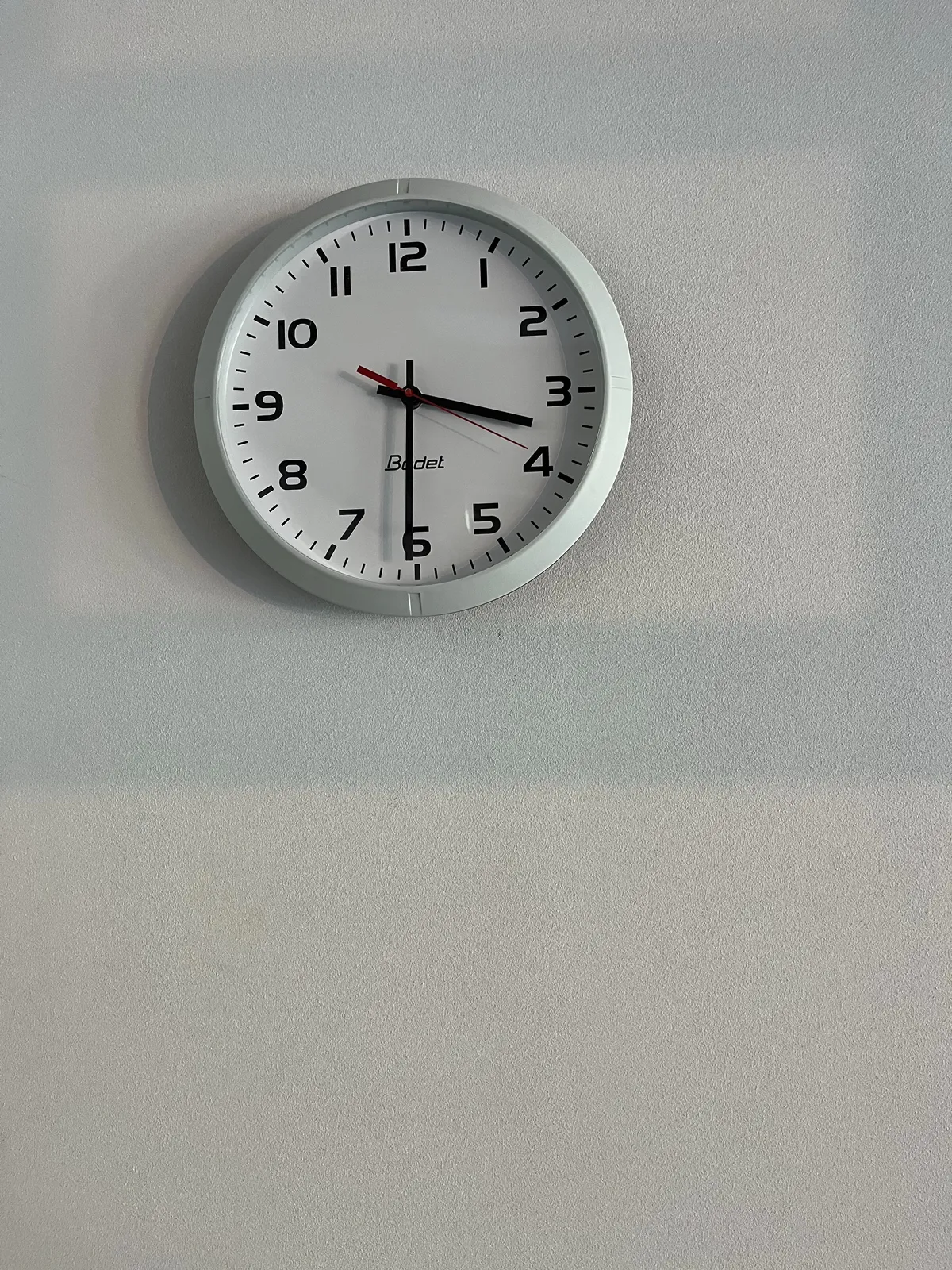 A plastic, white wall clock on a white wall, with a light shadow, cast by a window. The clock points at half past 3.