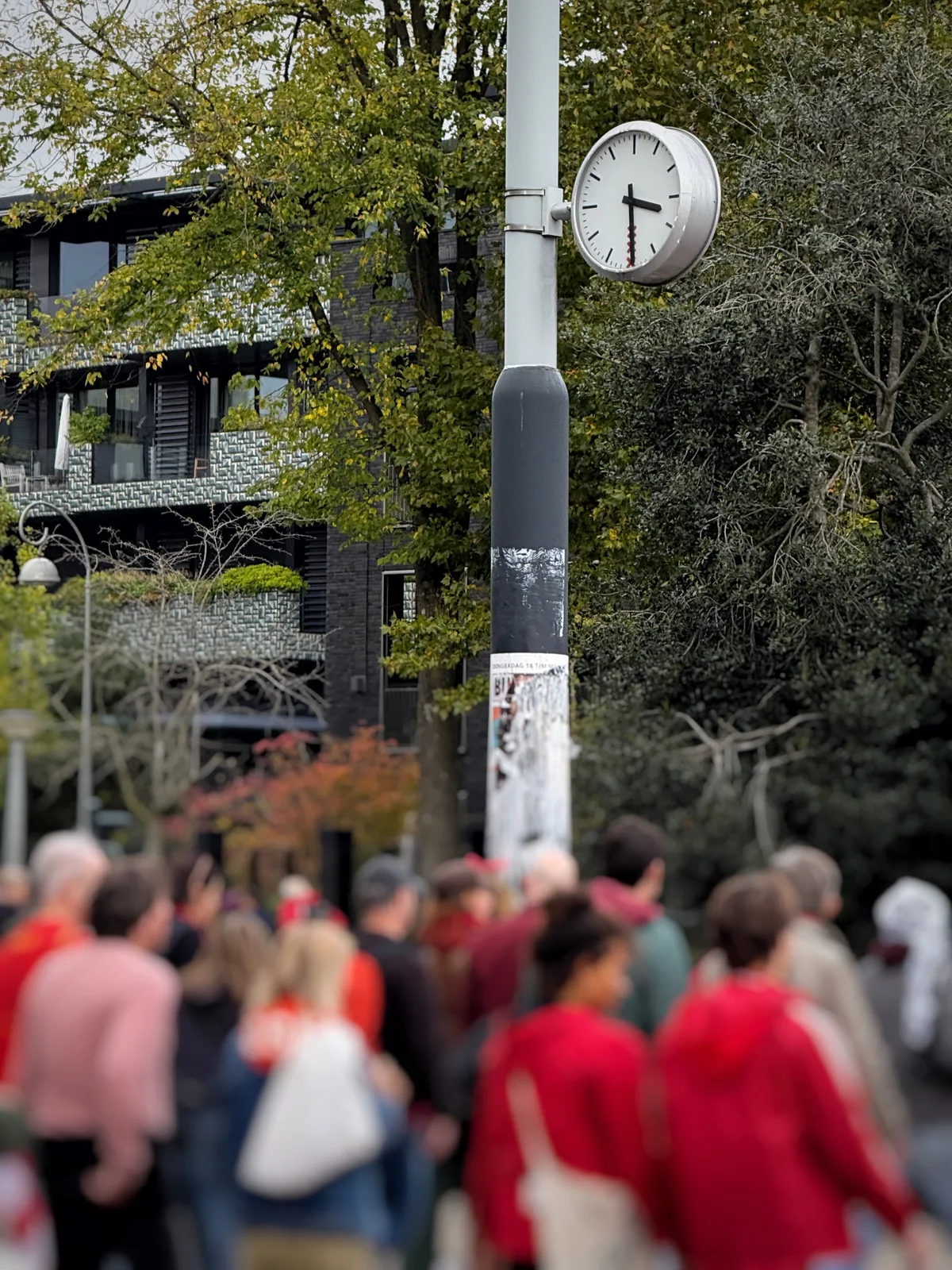 A blurred crowd of people below a clock on a pole. It points at half past three.