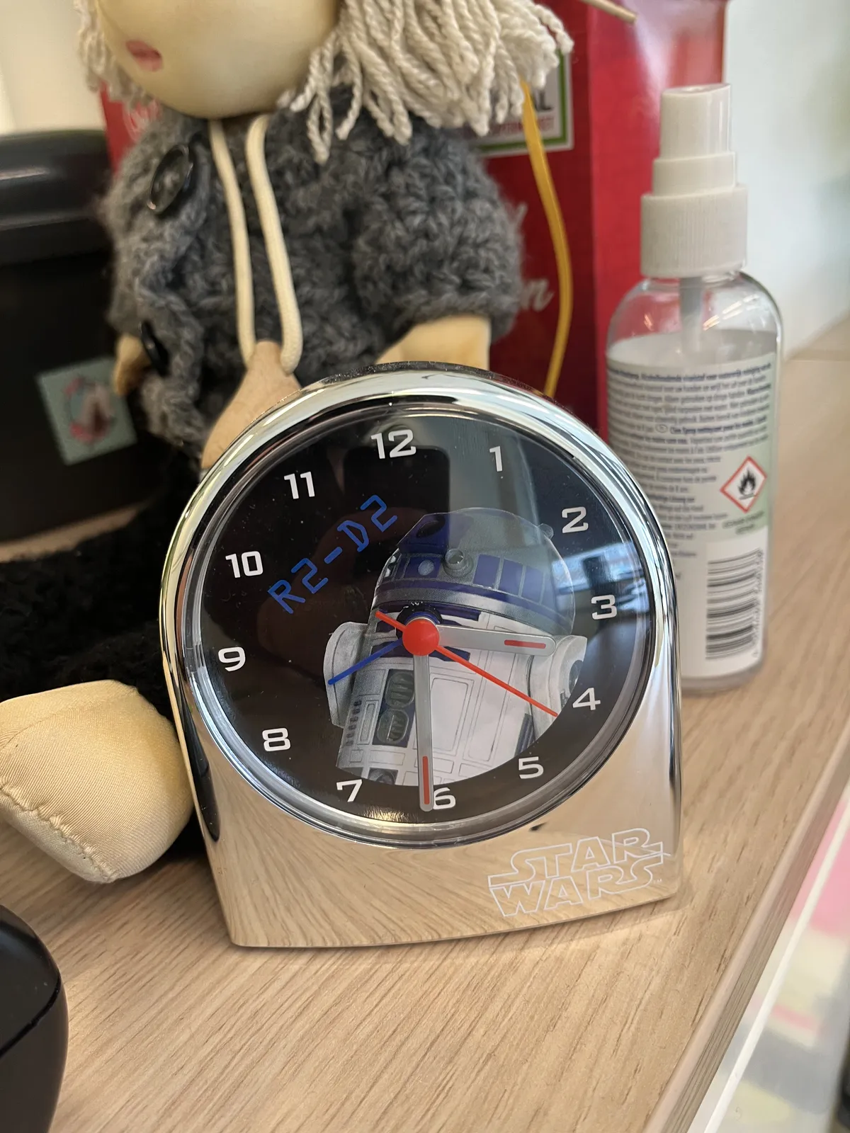 In front of a woollen doll there’s a star wars clock with r2d2 on its face. It points at 31 minutes past three.