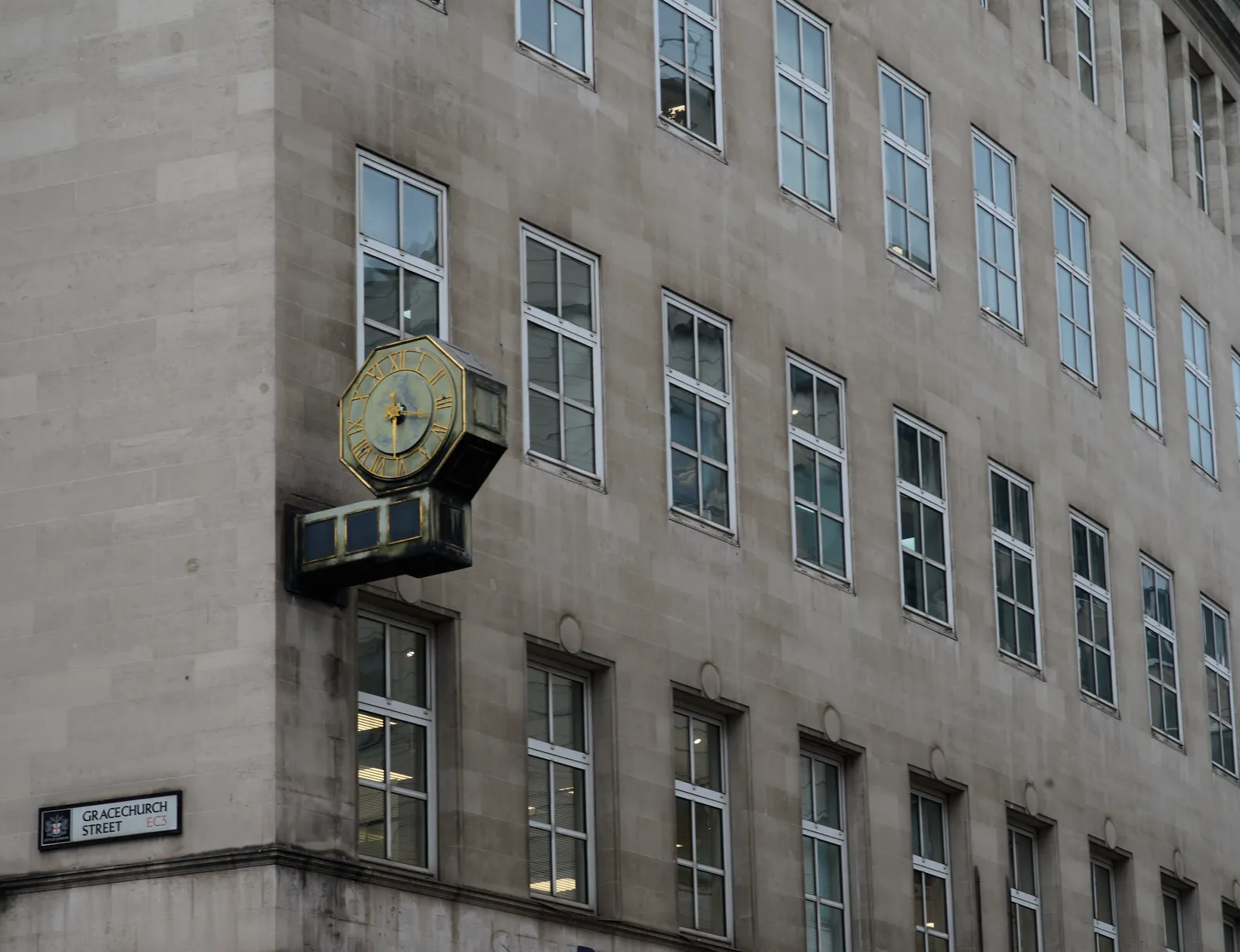 An old, brass clock, attached to a monumental facade around the corner of Gracechurch Street. The clock has oxidiced, so it is green/blue. It has golden hands and indicators and it points at 31 minutes past three.