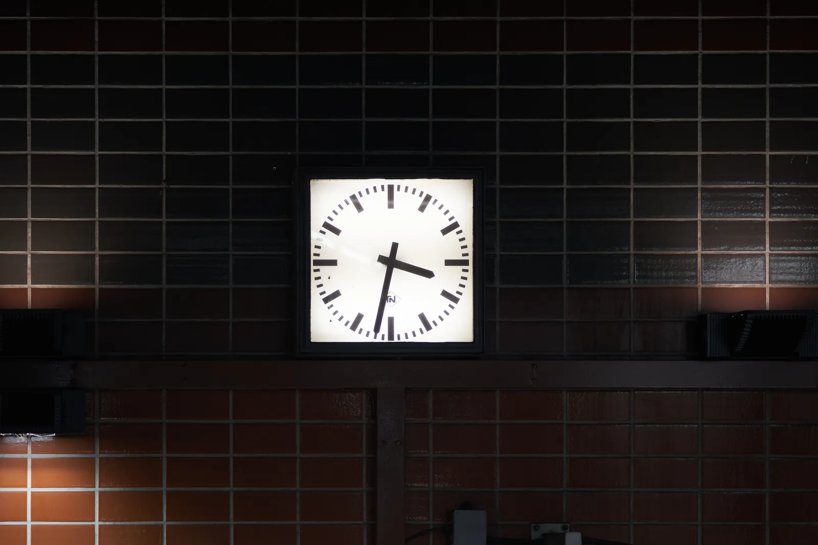 A clock on a tiled wall. The tiles are brown and grey. The clock is modernist and it points at 28 minutes to 4. 