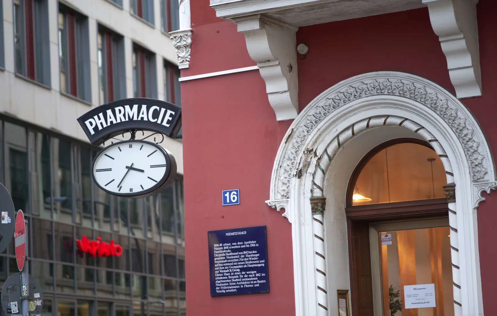 In the background there’s a grey modern office building, in the foreground there’s a more classic facade: a red wall, an ornamented white arched door, and a clock attached to the wall with the word Pharmacie printed above it. It points at 24 minutes to 4.
