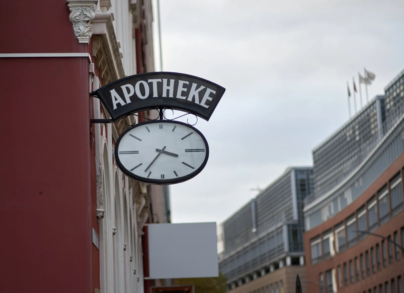 Rather boring office buildings in the background. In the foreground there’s a red wall with a clock attached to it with a sign that says Apotheke. The clock points at 23 minutes to 4.