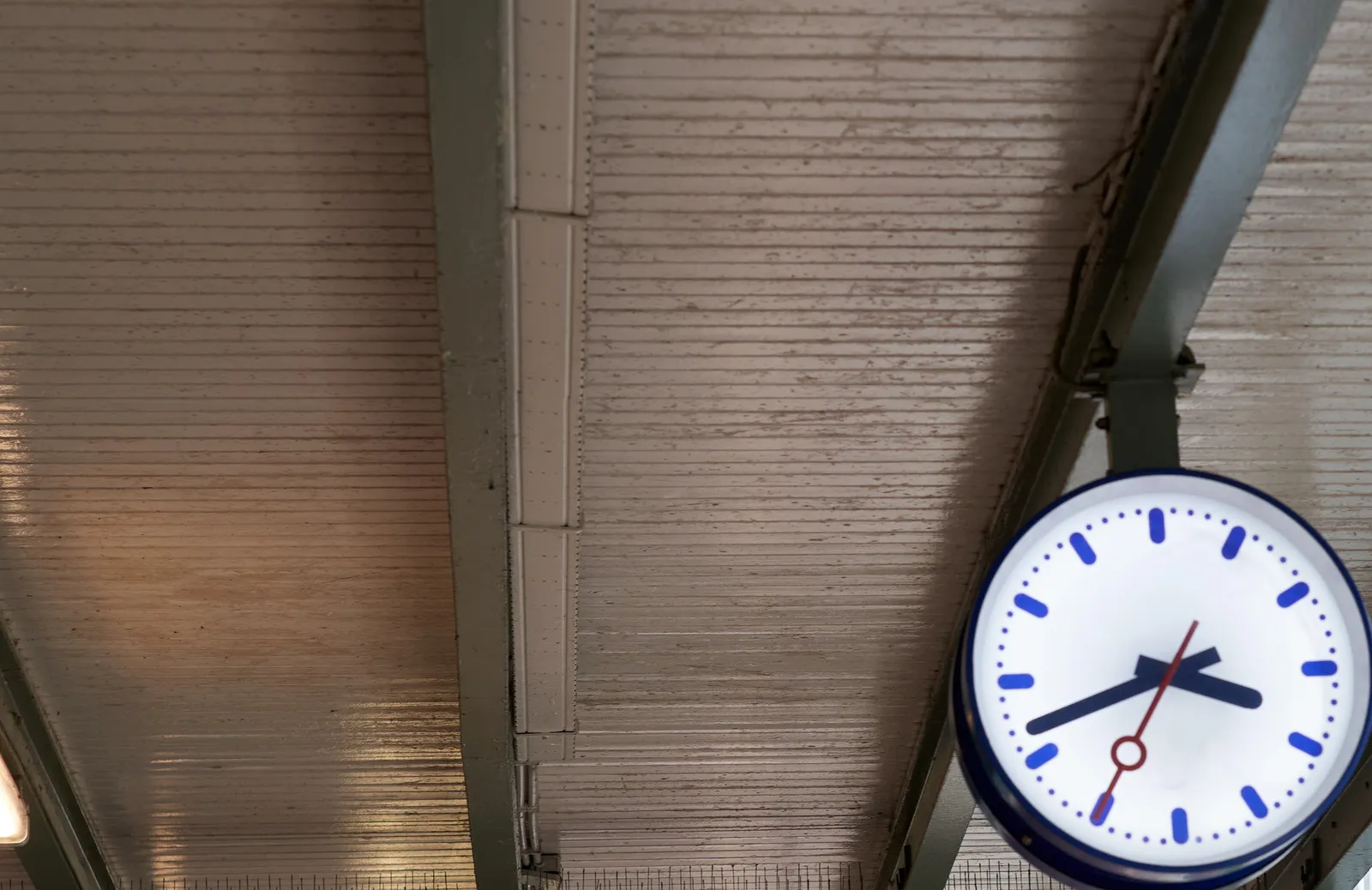 A clock, hanging from the ceiling in a train station. It points at 18 minutes to 4.