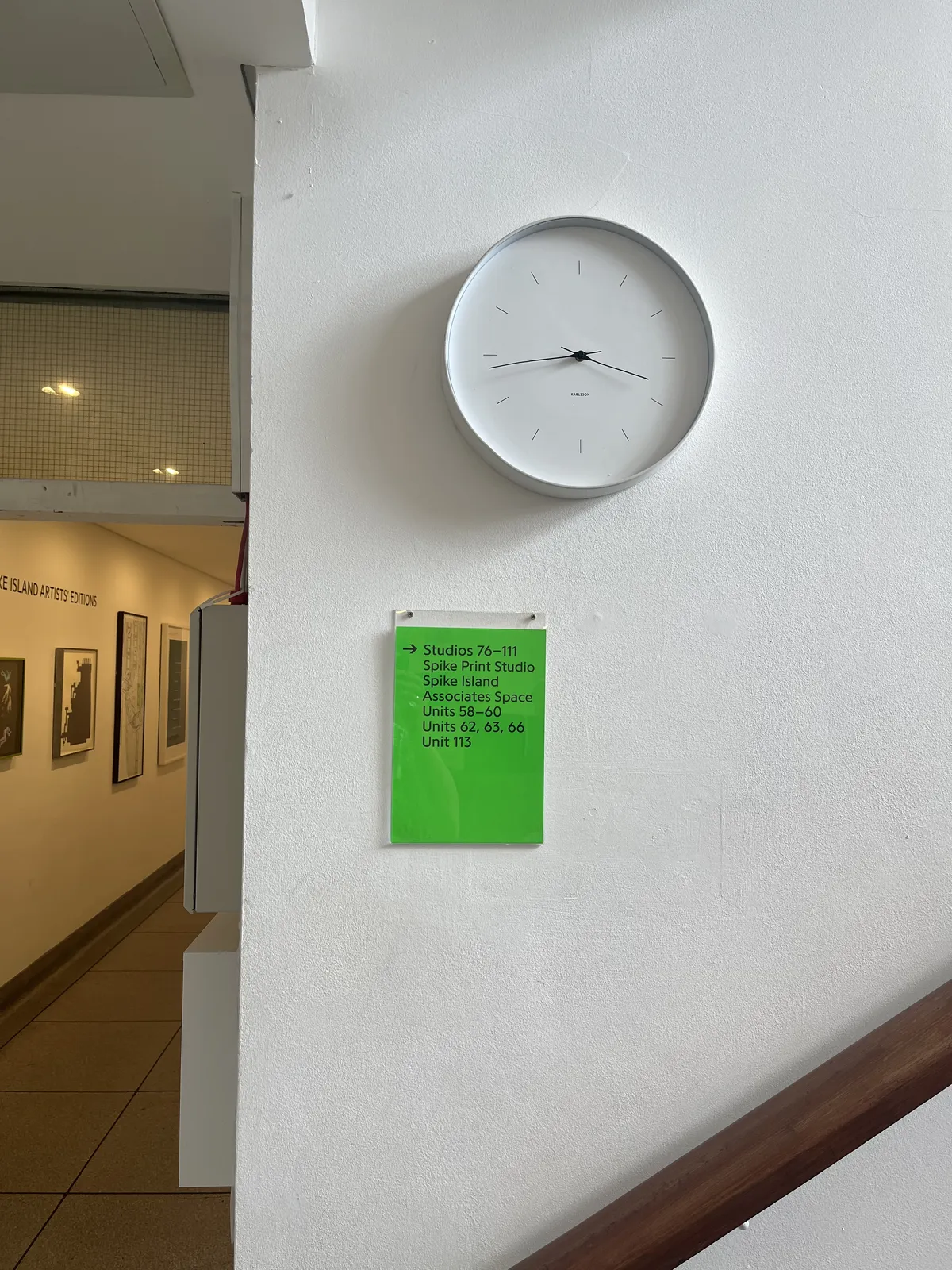 In the background there’s some art hanging in a hallway. On the wall in the foreground there’s a wooden handrail of stairs going up. There’s also a bright green piece of paper   with some information about studios and units. And there’s a minimalist wall clock that points at 16 minutes to four.