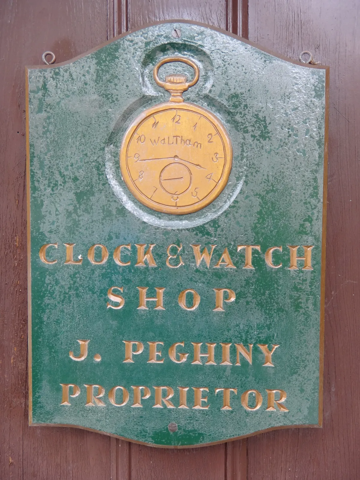 A green sign that says: Clock and Watch shop J Peghiny proprietor. There’s a pocket watch on this sign as well which points at approximately 16 minutes to 4.