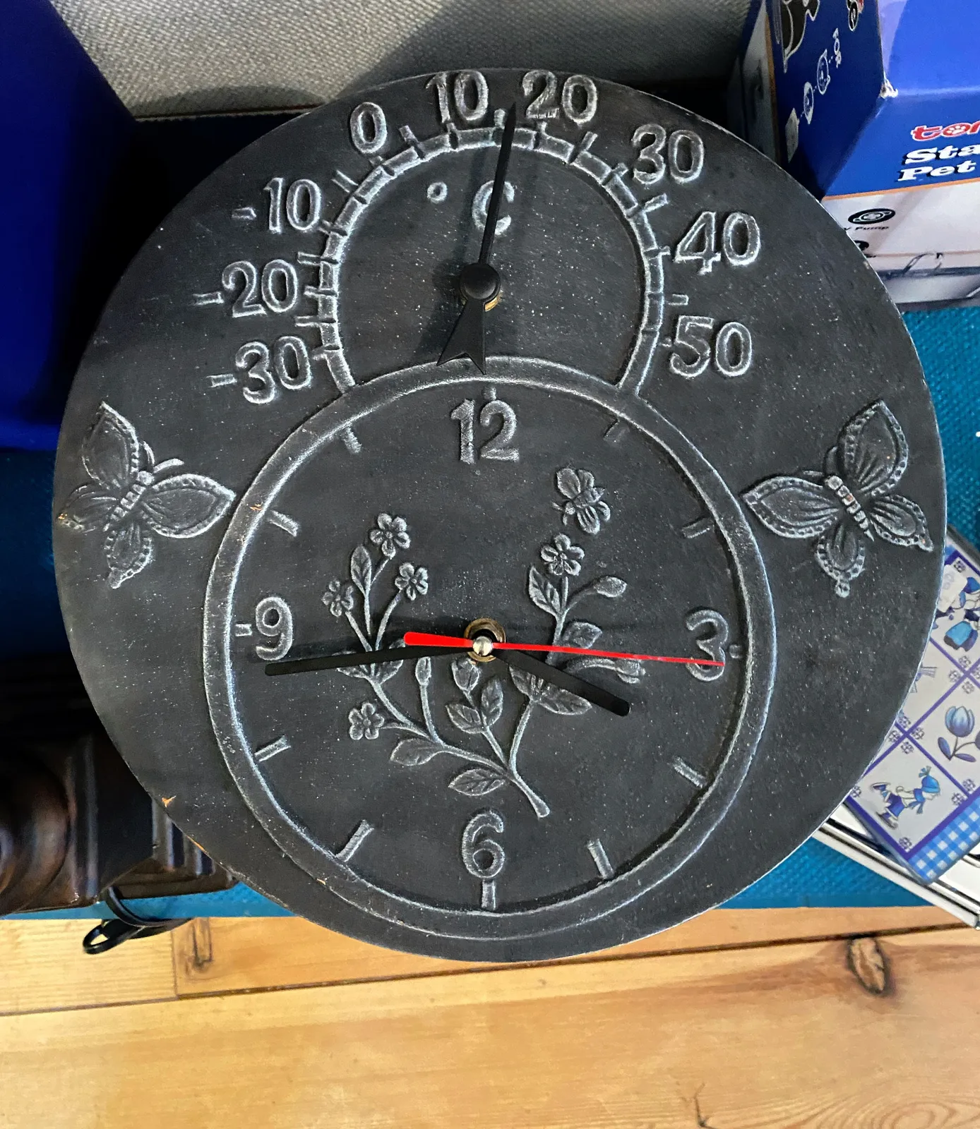 A black ceramic clock with flowers and butterflies on its face. There are two dials on this clock. The top dial shows the temperature, the other one points at 16 minutes to 4.
