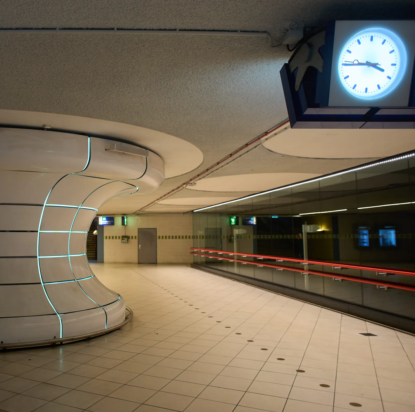 In the backgound there’s the empty hall of a Dutch train station, with a curvy work of art on the left. On the top right corner there’s a clock that points at a quarter to 4.