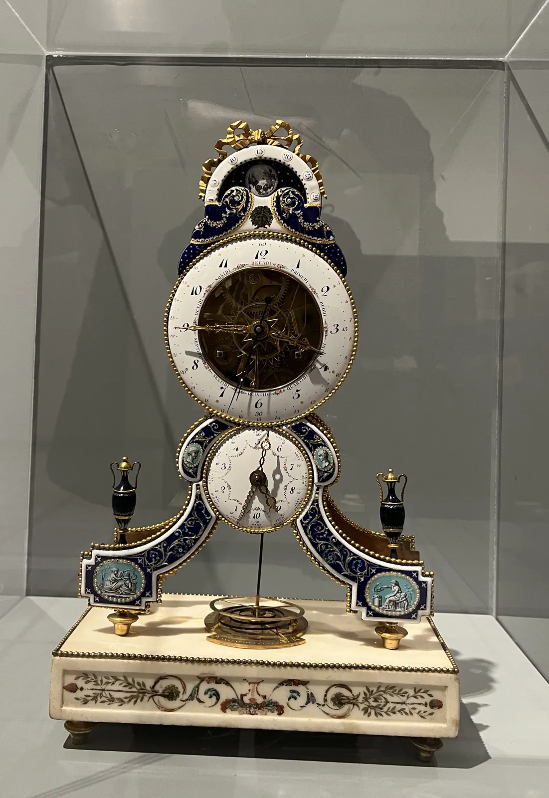 A rococo style mantel clock, richly ornamented. It has three dials. The one on the top probably shows the moon phases. The main one, in the middle, shows the time in the usual way, and at the moment it says it is a quater to four. The one on the bottom shows two things: it shows the months (and it indicates that it is almost April), and it shows the time in decimal numbers, but in an interestingly confusing way. The indicators of the 100 minutes start at the top, while the indicators of the 10 hours start at the bottom. So with quite some effort this decimal clock seems to point at approximately four minutes past nine, in decimal time. The two clocks are clearly not in sync.