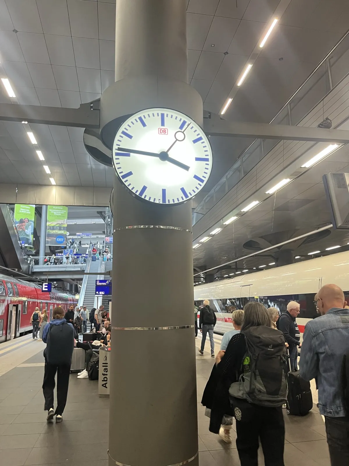 Many people, seen from the back, at a German train station. It is a German train station because the logo of the german railways is on the clock in the center of the picture. It points at 14 minutes to four.